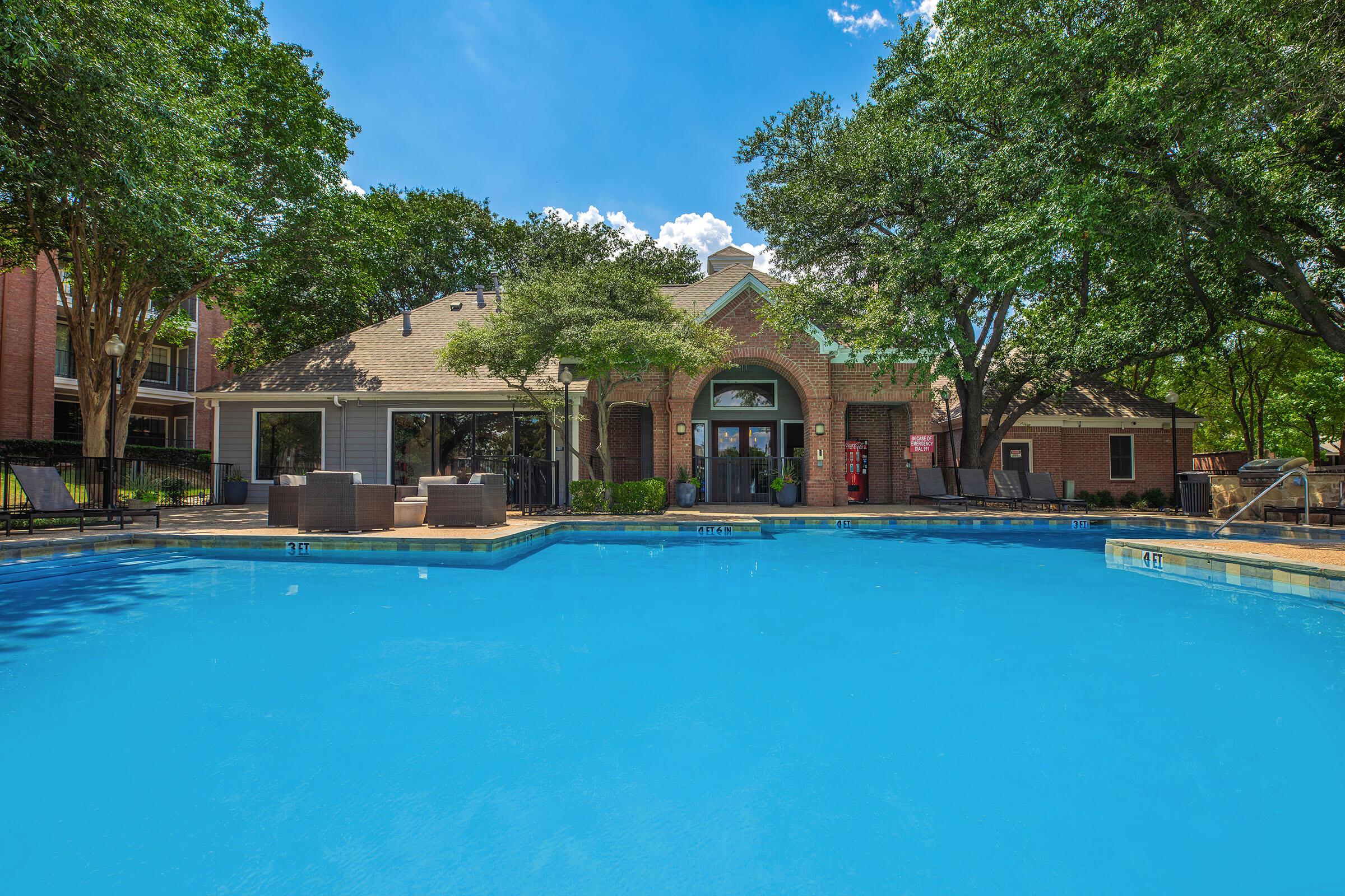 A bright and clear swimming pool surrounded by lounge chairs, with a building in the background featuring large windows and greenery. The sky is blue with a few clouds, and trees provide shade around the area, creating a relaxing atmosphere.