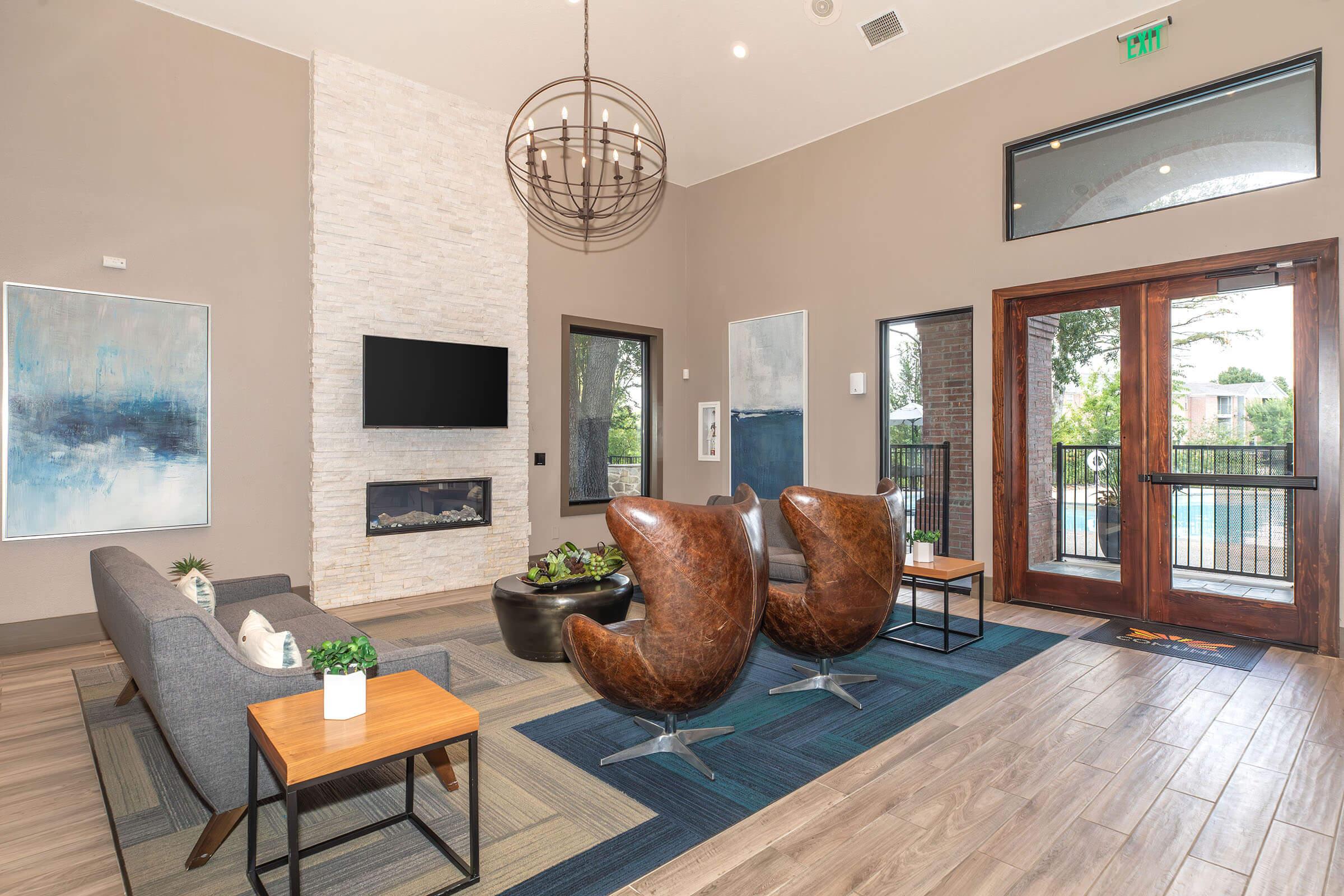 Modern lobby area featuring a stylish gray sofa, two brown leather chairs, a coffee table with plants, and a flat-screen TV mounted on a stone wall. Large windows provide natural light and a view of the outdoor pool area, complemented by contemporary artwork on the walls and an elegant chandelier overhead.