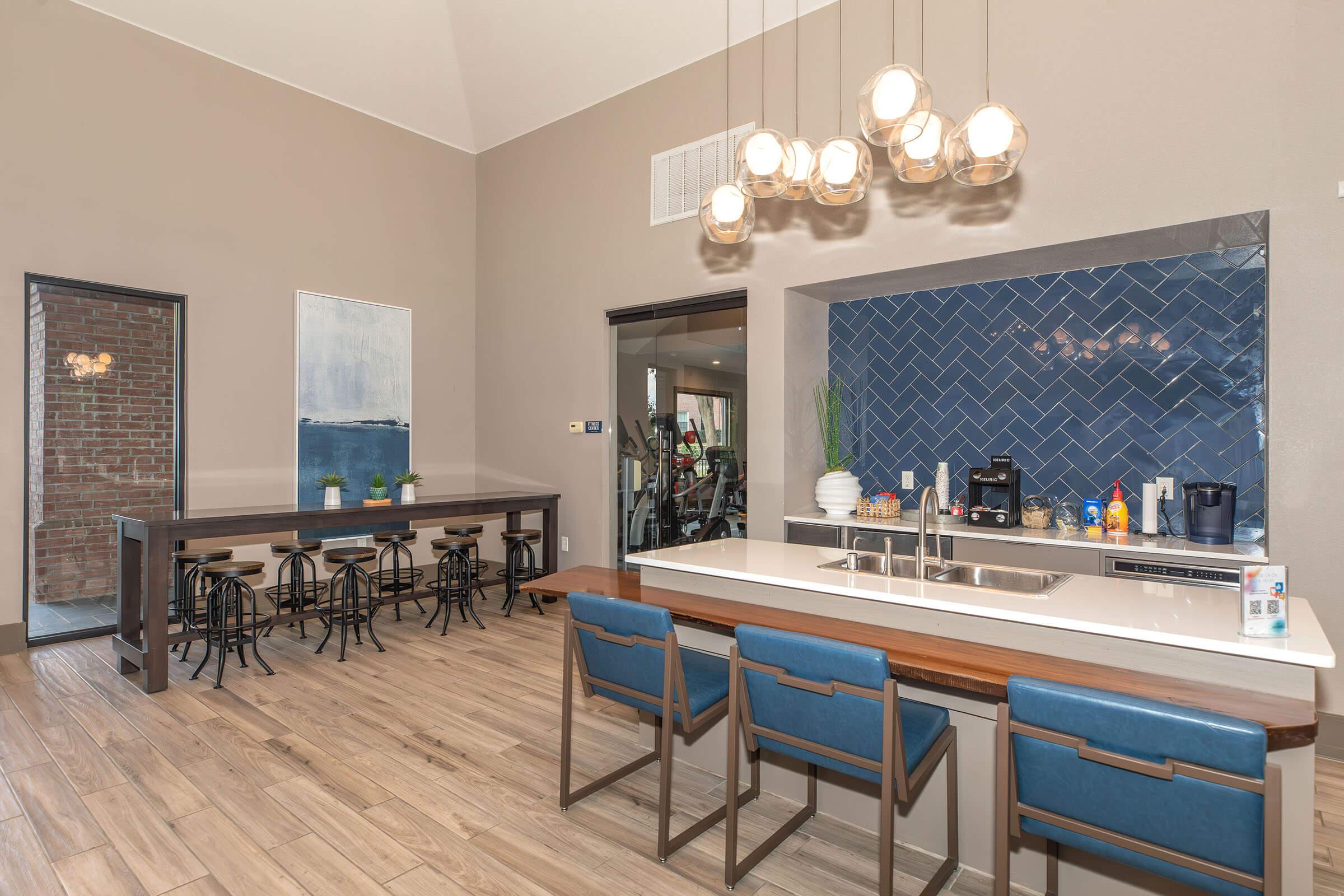 A modern kitchen and dining area featuring a long wooden table with black stools, a sleek white countertop with a blue backsplash, and contemporary light fixtures. The space is well-lit, with a painting on one wall and a glimpse of a fitness area visible through an open doorway.