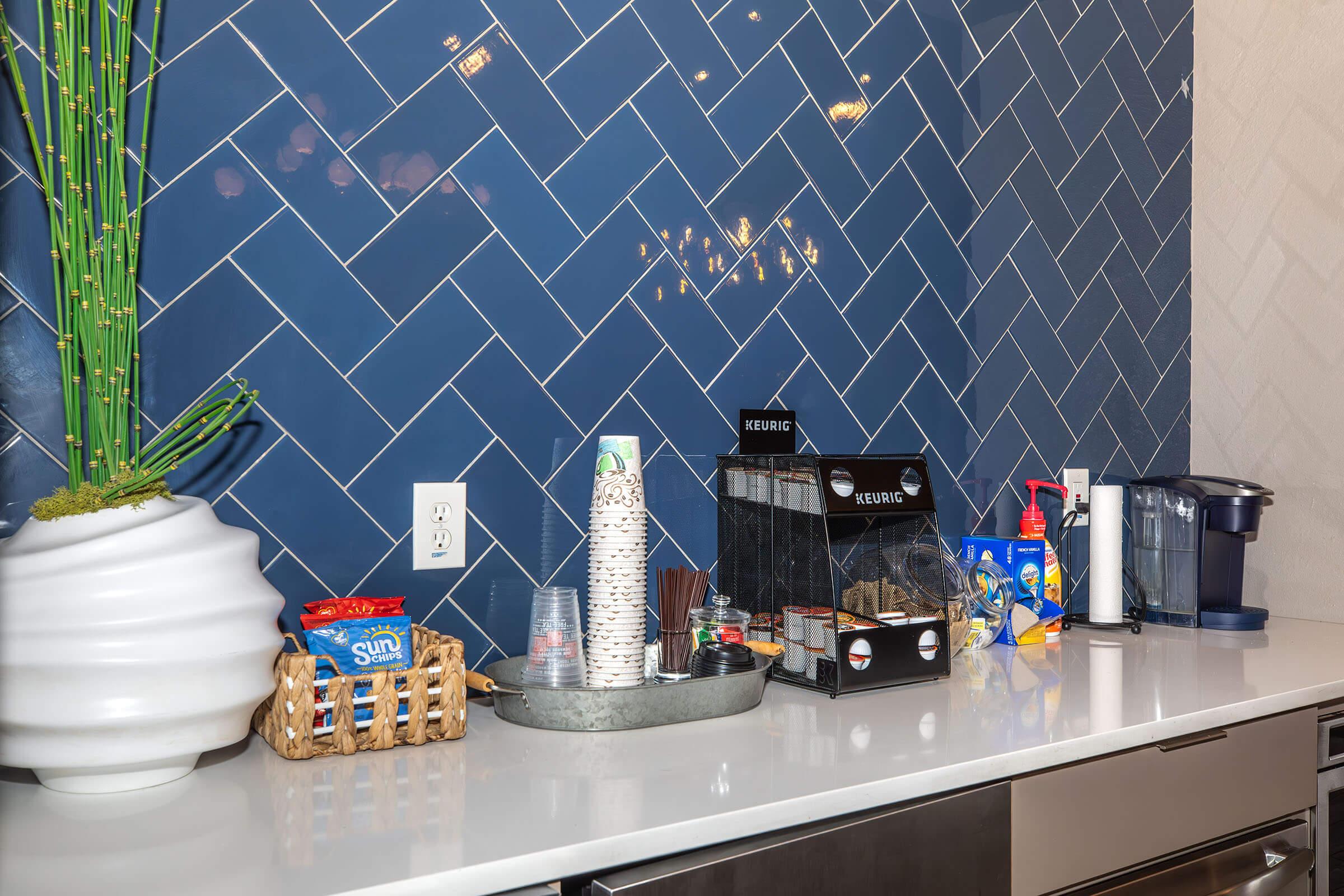 A modern kitchenette featuring a navy blue herringbone tiled wall. On the counter, a white ceramic vase with bamboo, a woven basket with snacks, a coffee maker, a cup dispenser, and various beverage supplies, including a pitcher and cups, arranged neatly on a sleek countertop.