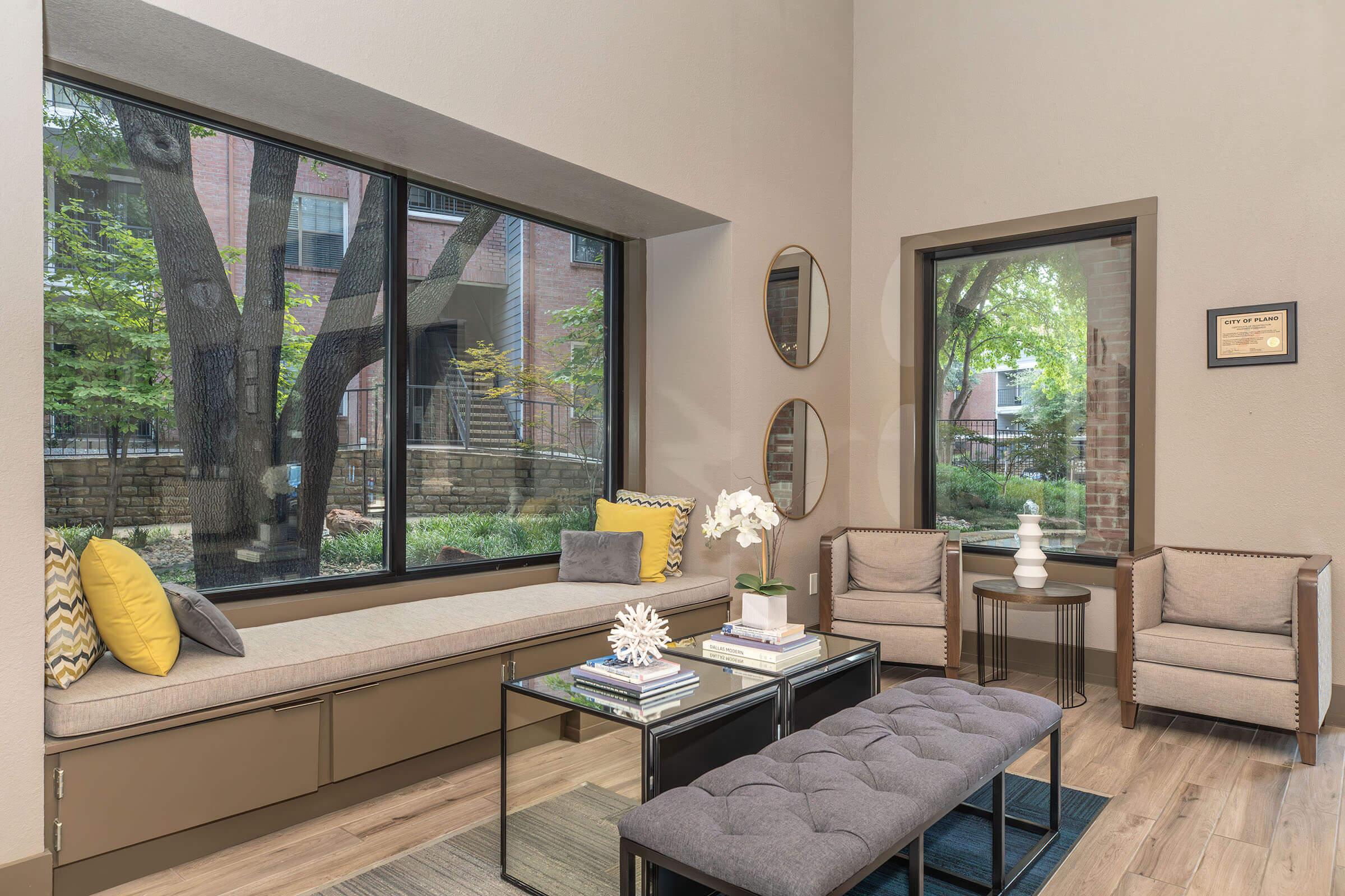 A cozy interior space featuring a large window with views of greenery outside. There is a built-in bench with yellow and gray cushions, a coffee table with decorative items, and two modern armchairs. The walls are neutral-colored, and there are circular mirrors and a framed certificate on display.
