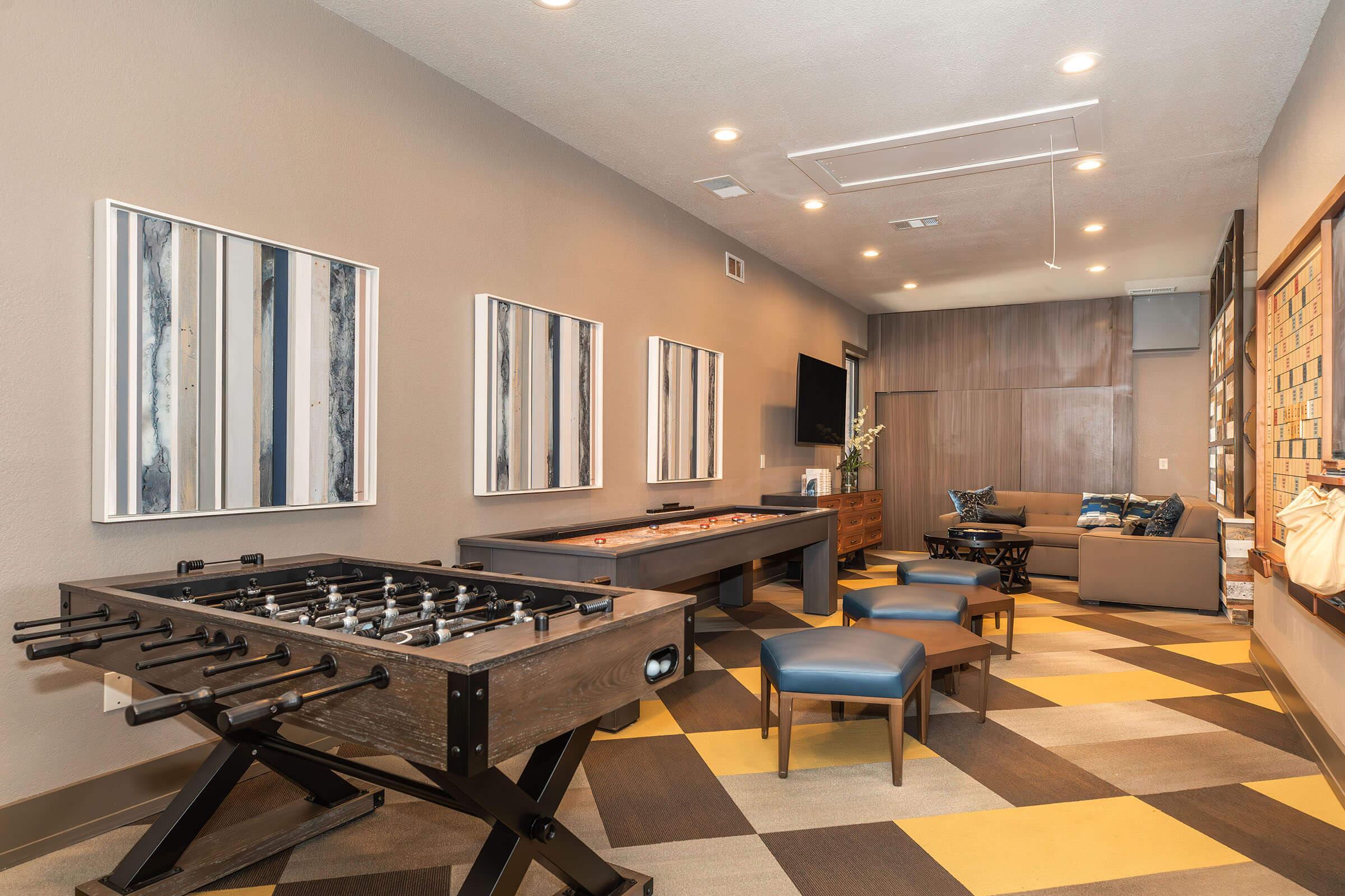 A modern game room featuring a foosball table, an air hockey table, and blue patterned chairs. The walls are adorned with abstract artwork, and there is a comfortable seating area with a couch. The floor has a geometric pattern in yellow and gray, creating a vibrant and inviting atmosphere.