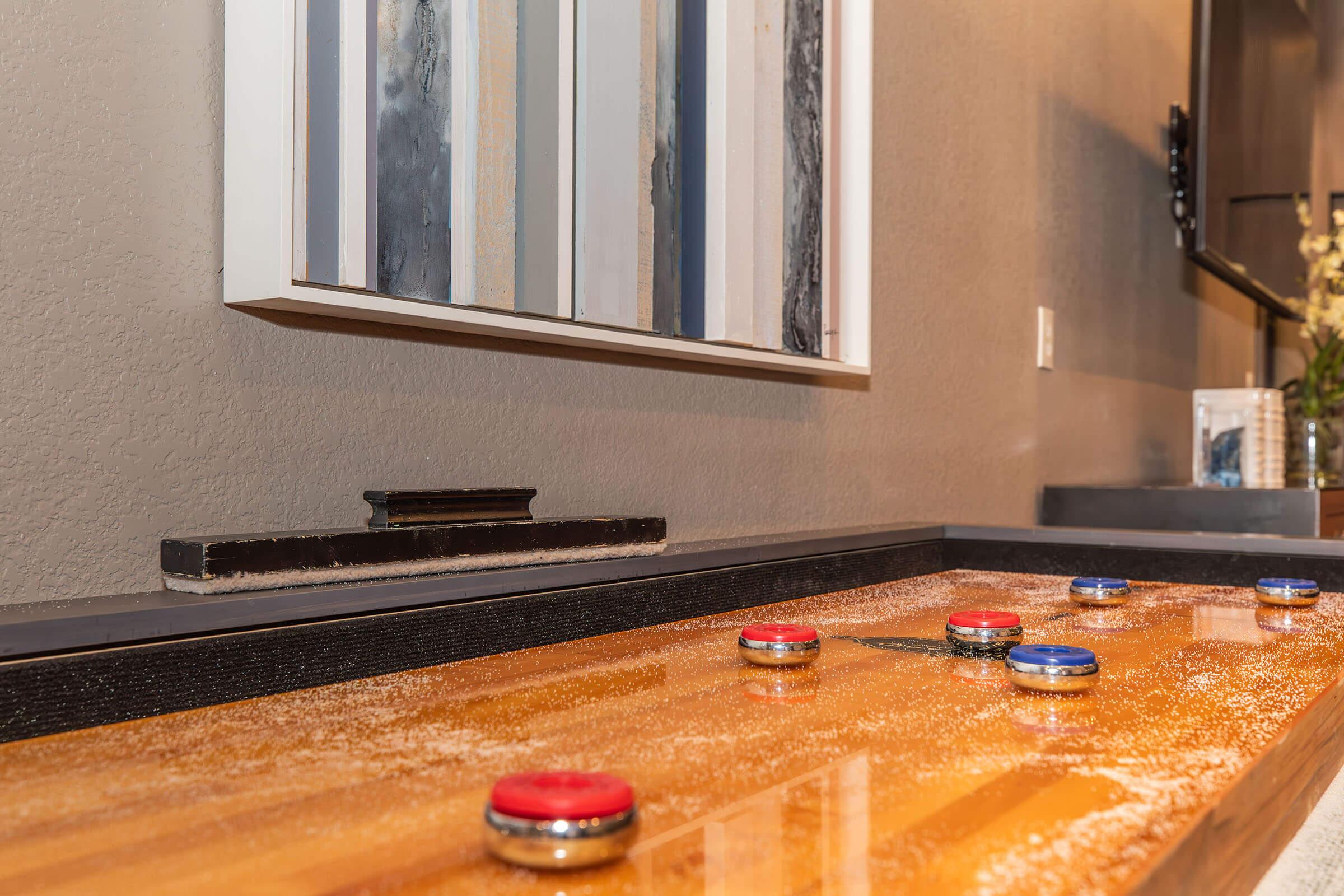 A shuffleboard table with red and blue pucks positioned on a wooden surface, lightly dusted with salt. A modern art piece hangs on the wall behind, featuring abstract patterns in muted colors. The setting has a contemporary aesthetic, with a flat-screen TV partially visible in the background.
