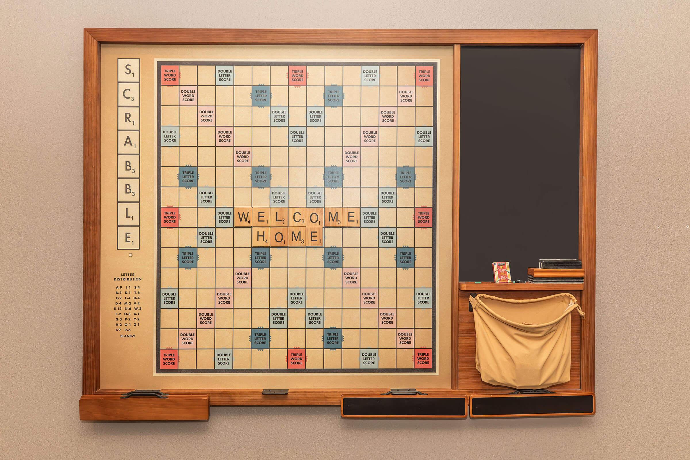 A wooden wall-mounted Scrabble board displaying the words "WELCOME HOME" in the center. The board features a classic grid layout with blue and red squares, tiles stored in a pocket on the side, and a chalkboard section for notes or scoring.