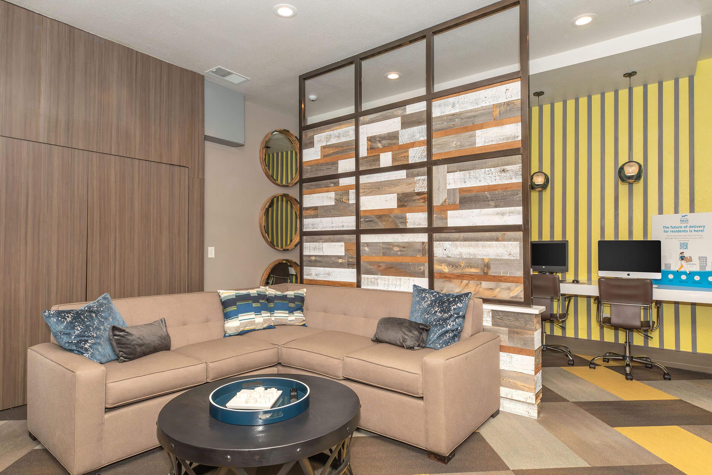 A stylish lounge area featuring a beige sectional sofa with decorative pillows, a round coffee table, and a partition with rustic wood accents. In the background, there are two computer stations against a bright yellow and striped wall, along with circular mirrors reflecting the contemporary design.