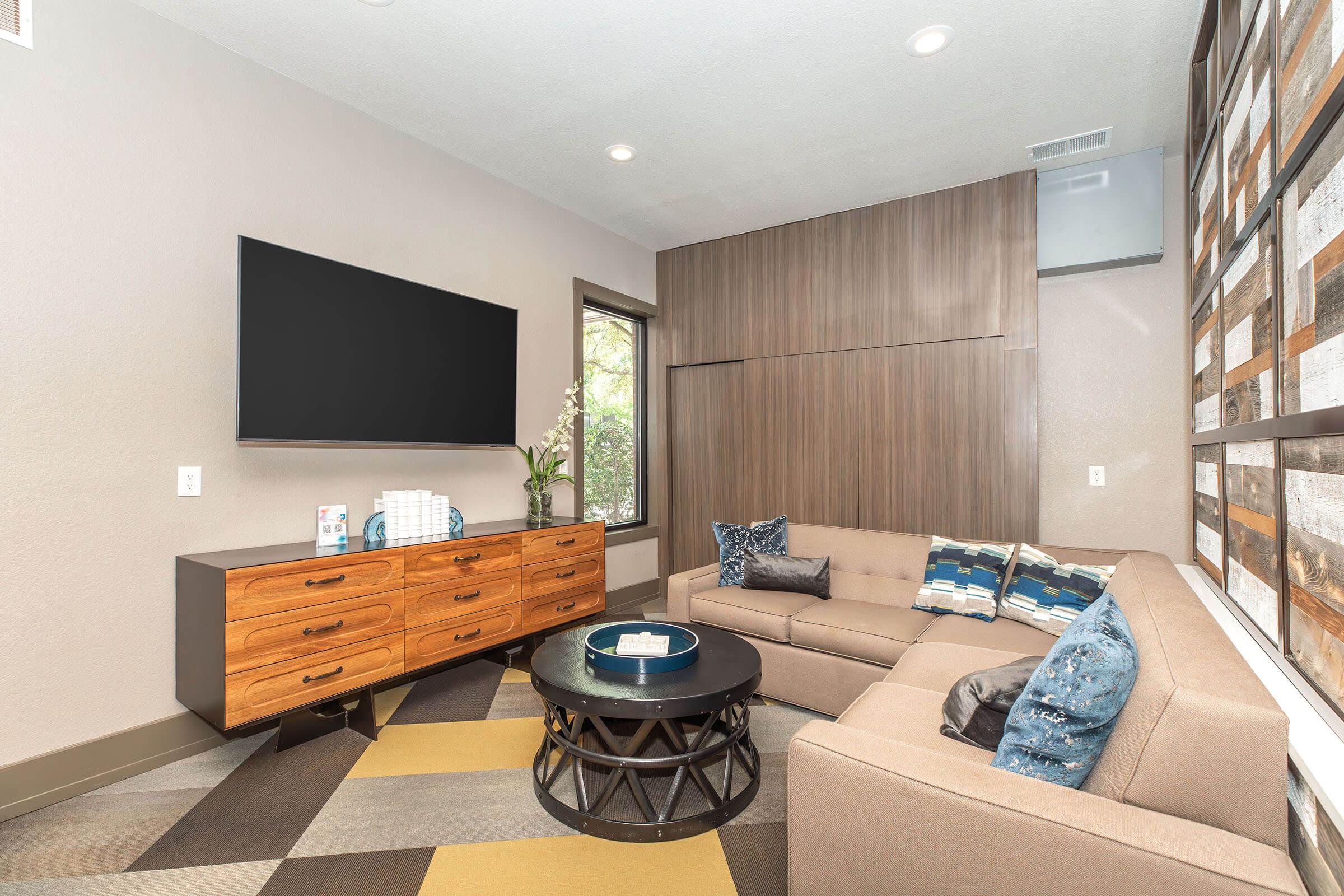 Modern living room featuring a beige sectional sofa with decorative blue pillows, a round black coffee table, and a stylish wooden TV stand with a flat-screen TV mounted on the wall. The walls are painted in neutral tones, and there is a decorative panel with a wood pattern. Bright natural light comes through the window.