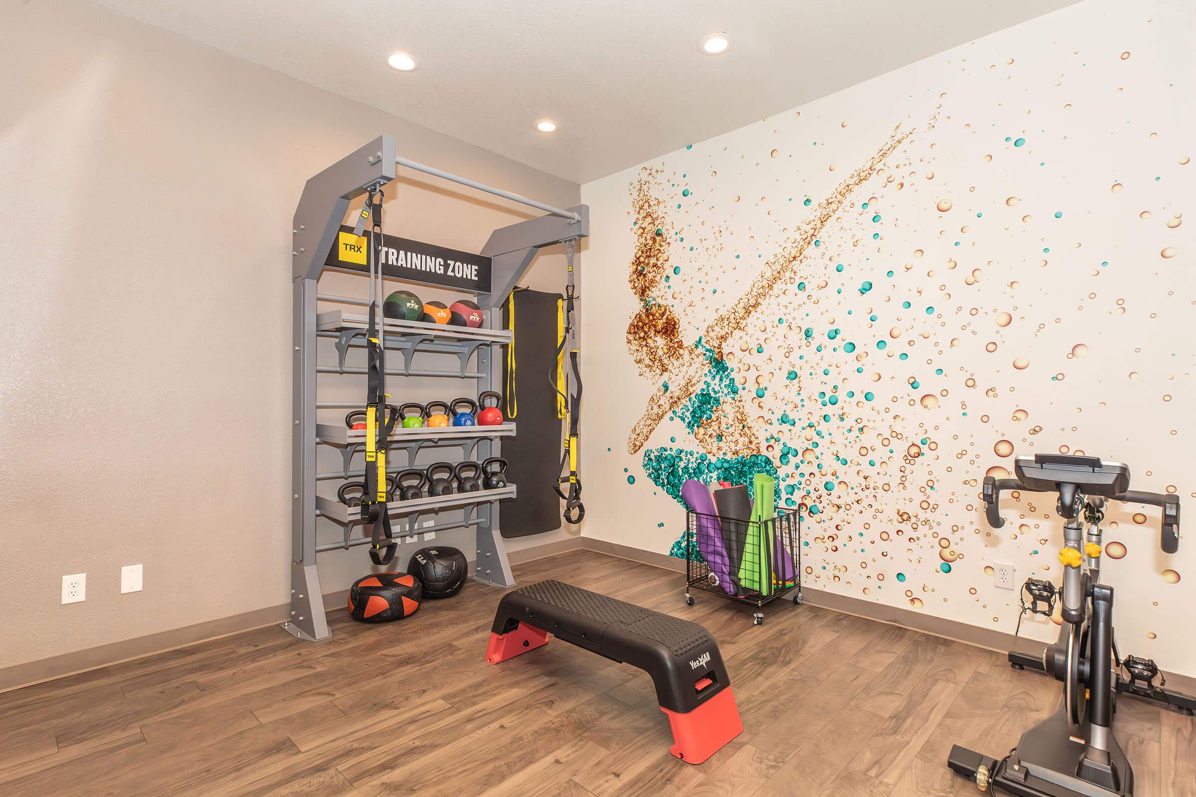 A modern training room featuring a multi-use exercise rack with kettlebells and suspension straps, a bench for workouts, a stationary bike, and a colorful wall mural with bubbles and a silhouette of a person in motion. The space is well-lit with overhead lights and has a sleek, contemporary design.