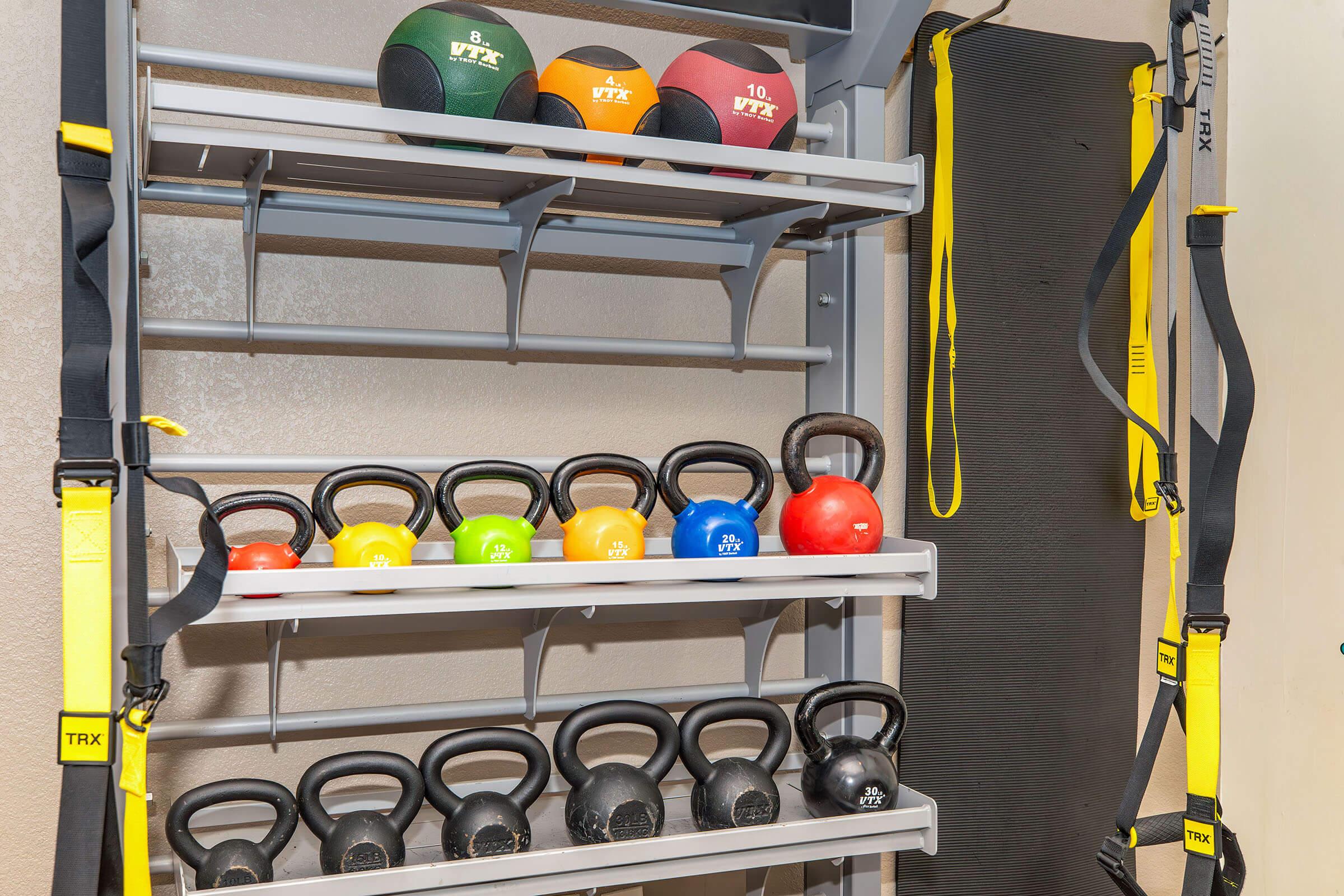 A home gym setup featuring a vertical rack holding various kettlebells in multiple colors, including red, yellow, and black. TRX suspension straps hang on the side, alongside a gray exercise mat on the wall. The environment appears well-lit and designed for fitness activities.