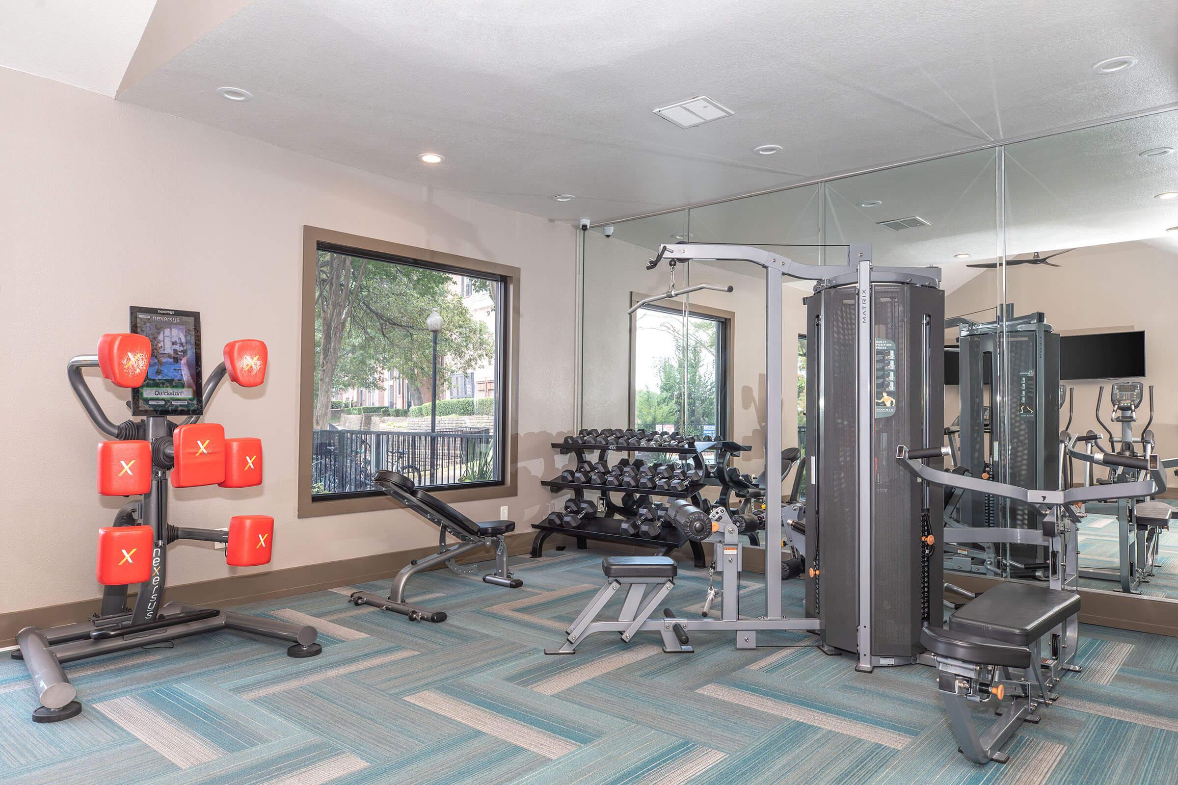 A modern gym interior featuring various workout equipment, including a weight bench, dumbbells, and a multi-station exercise machine. Large windows allow natural light, and a mirrored wall reflects the space. The flooring has a blue and green striped pattern.