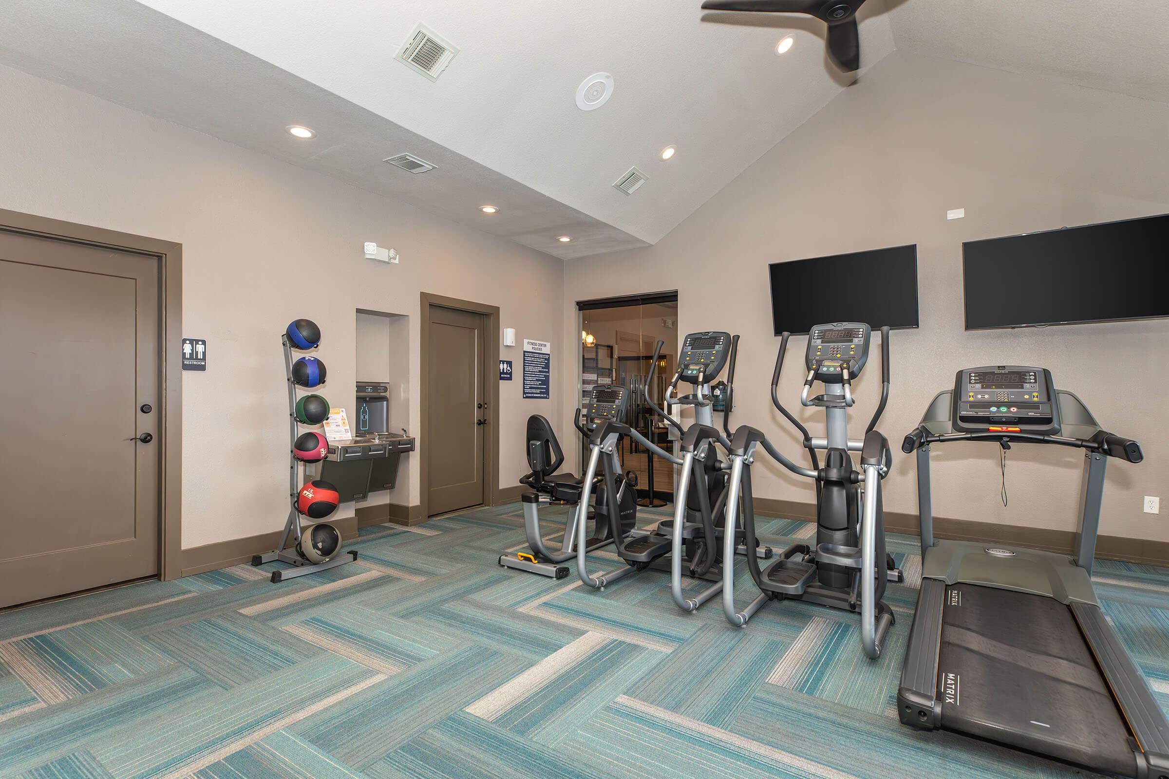 A modern gym interior featuring exercise equipment including treadmills, ellipticals, and a stationary bike. There are two wall-mounted TVs, a wall-mounted weight rack with colored medicine balls, and a chair in the corner. The flooring has a blue and green pattern, and the walls are painted light gray.