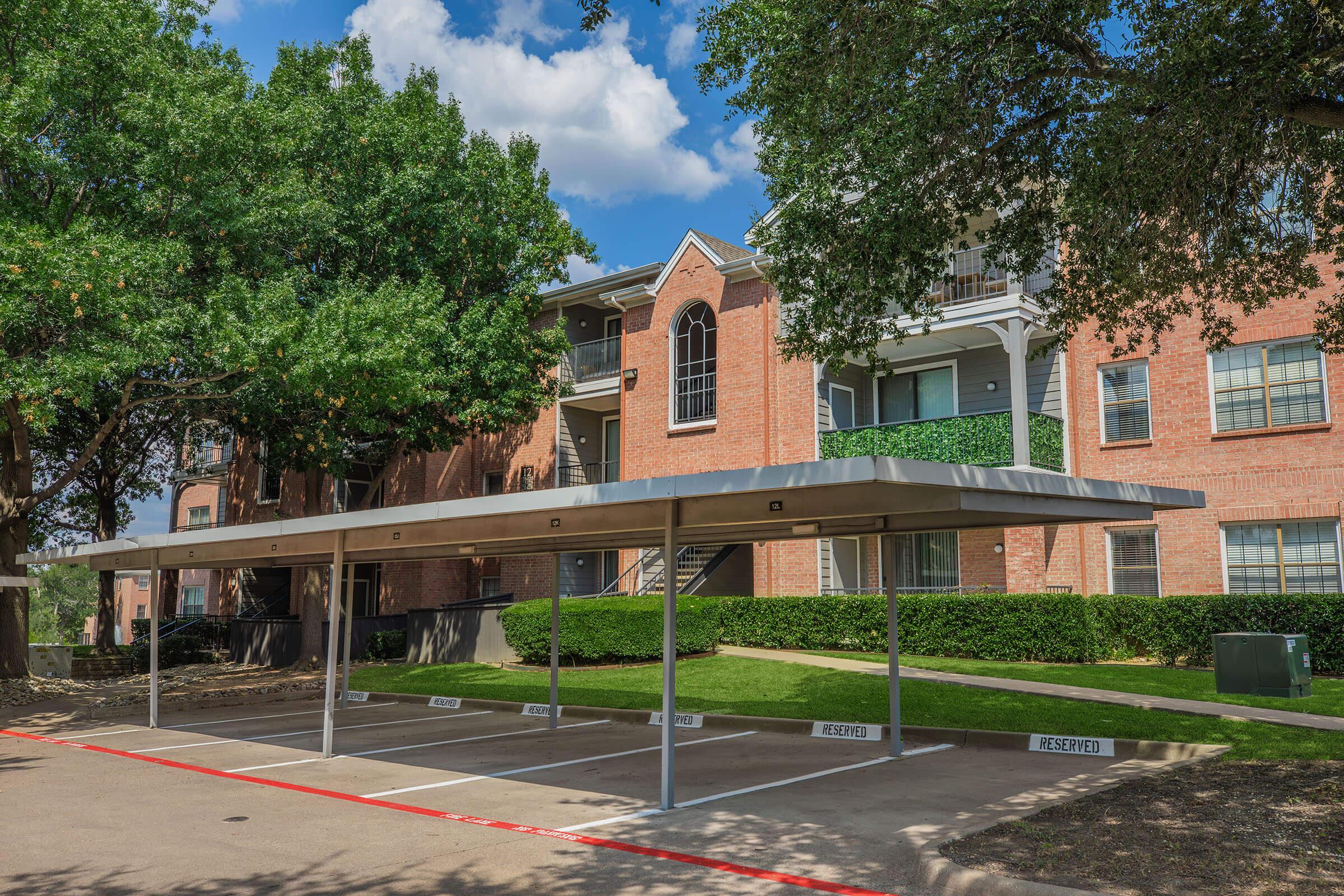 A parking lot with several reserved spaces, featuring a large carport. In the background, a multi-story brick apartment building is surrounded by trees and greenery. The sky is partly cloudy, adding a bright atmosphere to the scene.