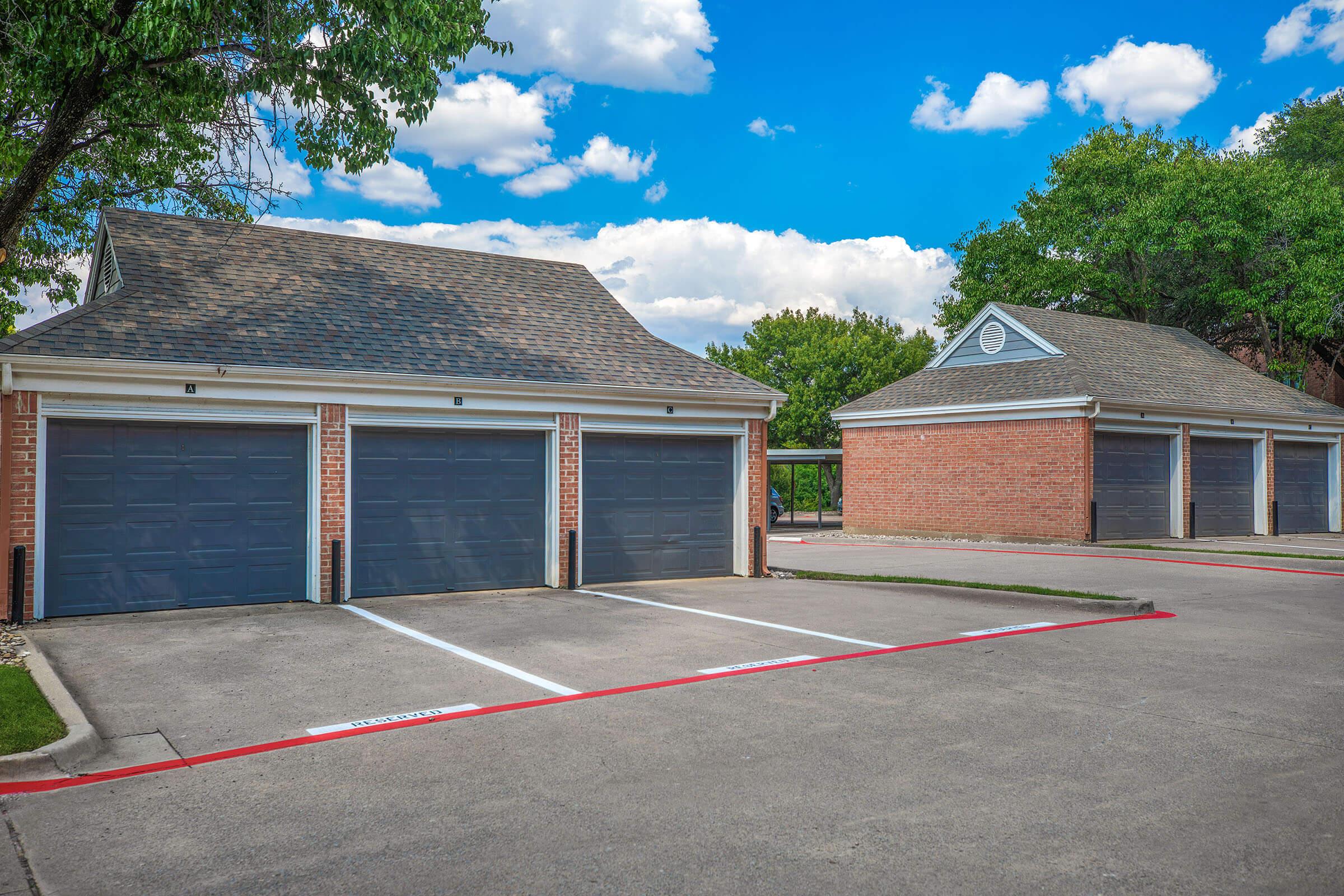 A parking area featuring two garage buildings with brown garage doors, surrounded by green trees and a blue sky with clouds. The asphalt surface has marked parking spots, and the overall environment appears well-maintained and spacious.
