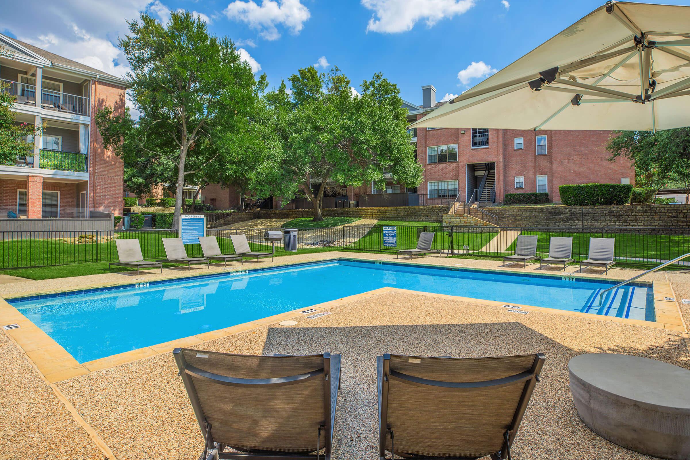 A clear blue swimming pool surrounded by lounge chairs, with green lawns and trees in the background. Nearby, there are apartment buildings, creating a relaxed outdoor setting. A shaded area is provided by a large umbrella. The sky is partly cloudy, adding to the inviting atmosphere.