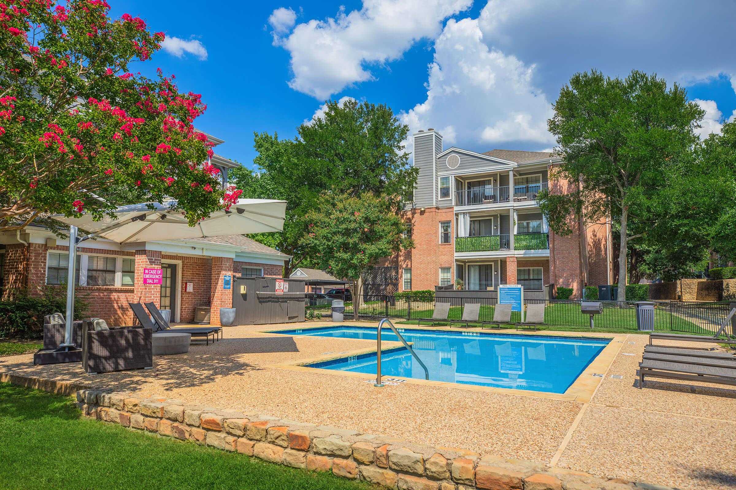 A sunny pool area at an apartment complex, featuring a rectangular swimming pool with a ladder, surrounded by lounge chairs. Lush green grass and trees provide a pleasant environment, while a shaded seating area with an umbrella is visible nearby, along with brick buildings in the background.