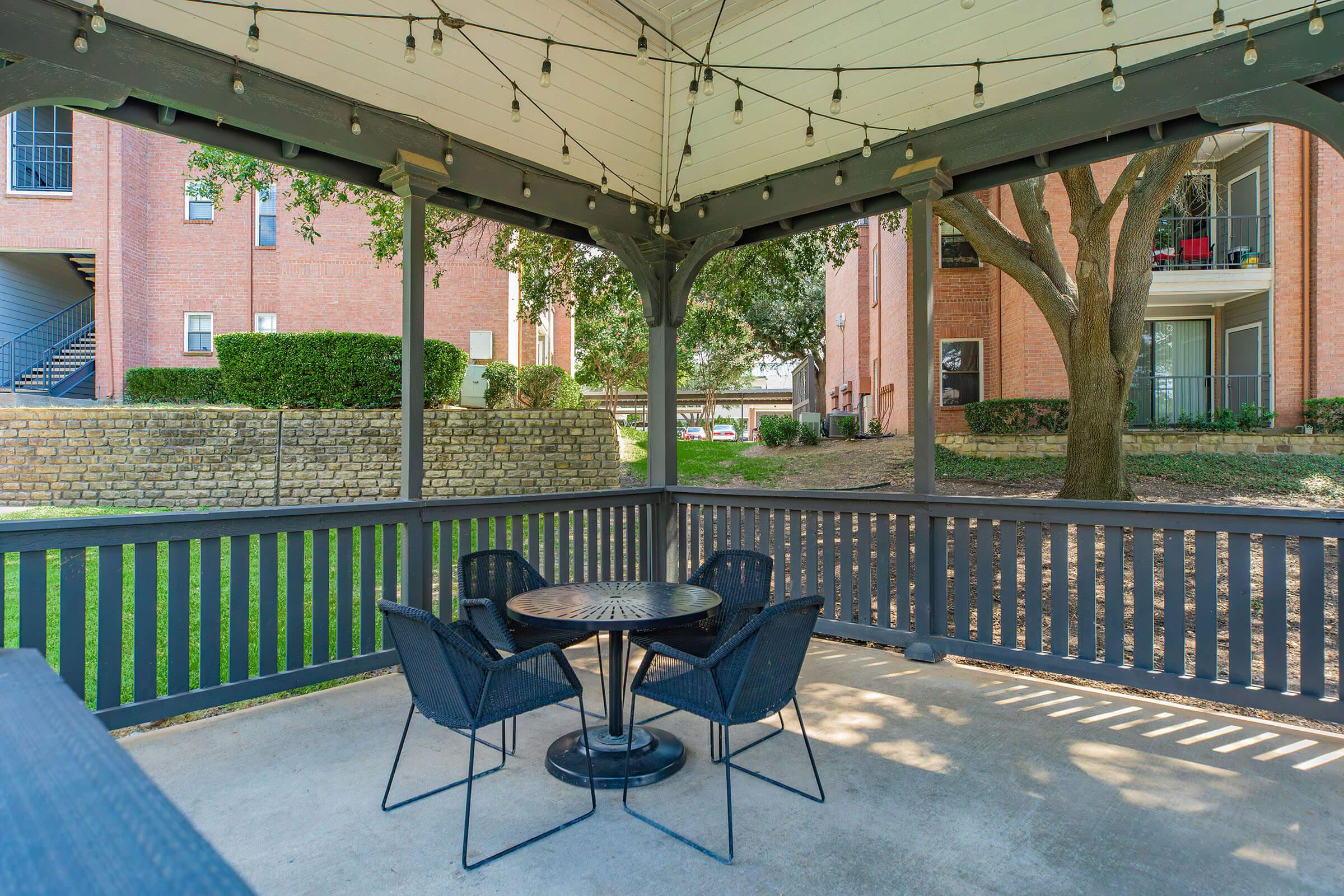 A shaded gazebo with a round table and four black chairs, surrounded by grassy areas and trees. The structure features string lights hanging from the ceiling, with brick buildings visible in the background.