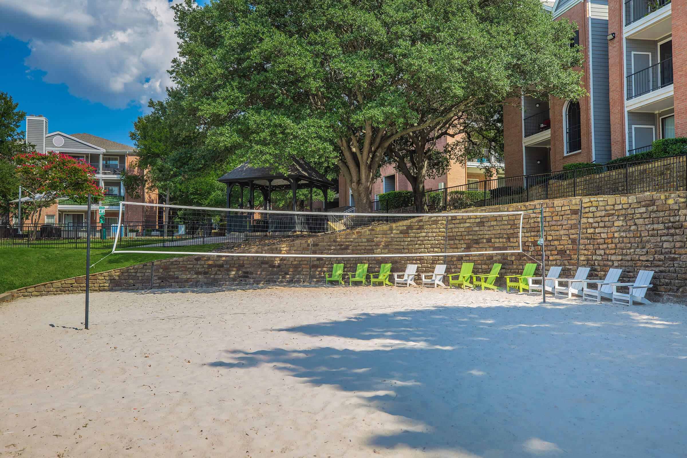 Sand volleyball court with a net set up, surrounded by green and white lounge chairs. In the background, there are trees and apartment buildings, with a gazebo nearby. The scene is bright and sunny, creating a relaxed recreational atmosphere.
