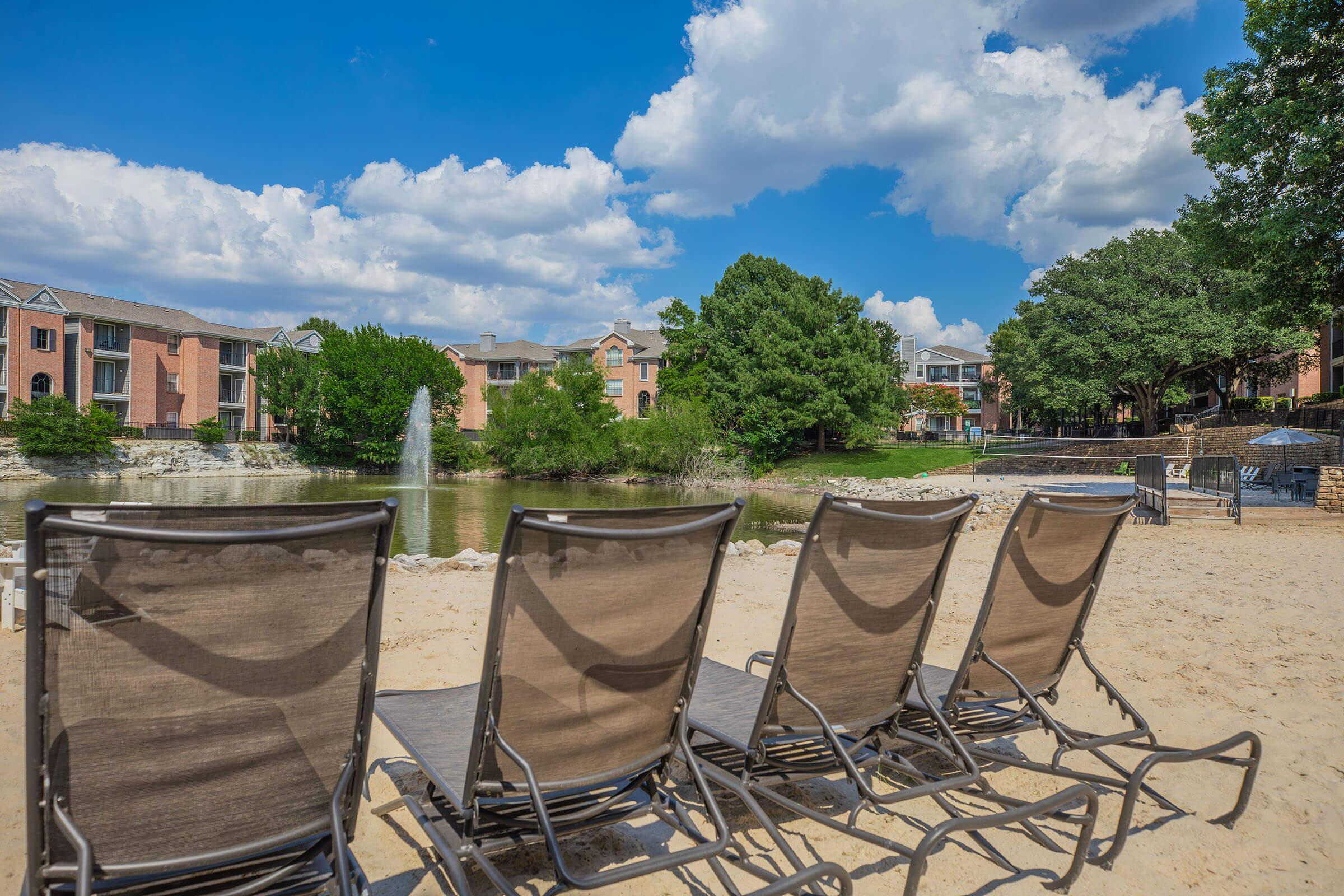 A scenic view of a sandy area by a calm lake, featuring four lounge chairs facing the water. In the background, green trees and residential buildings are visible under a bright blue sky with fluffy white clouds. A small fountain adds a serene touch to the setting.