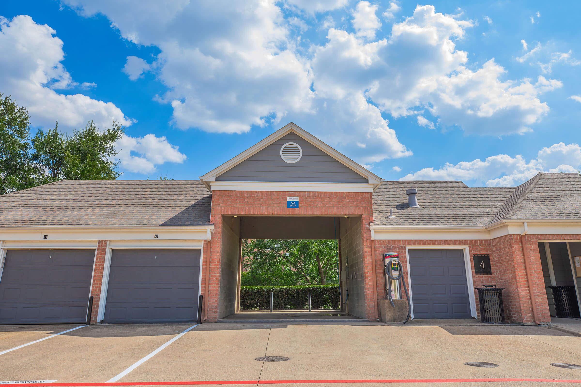 A brick building with garage doors, featuring a central entrance under a peaked roof. Two garage doors are visible on the left, one on the right. The area is well-lit with blue skies and scattered white clouds in the background. A telephone booth is positioned near the entrance.