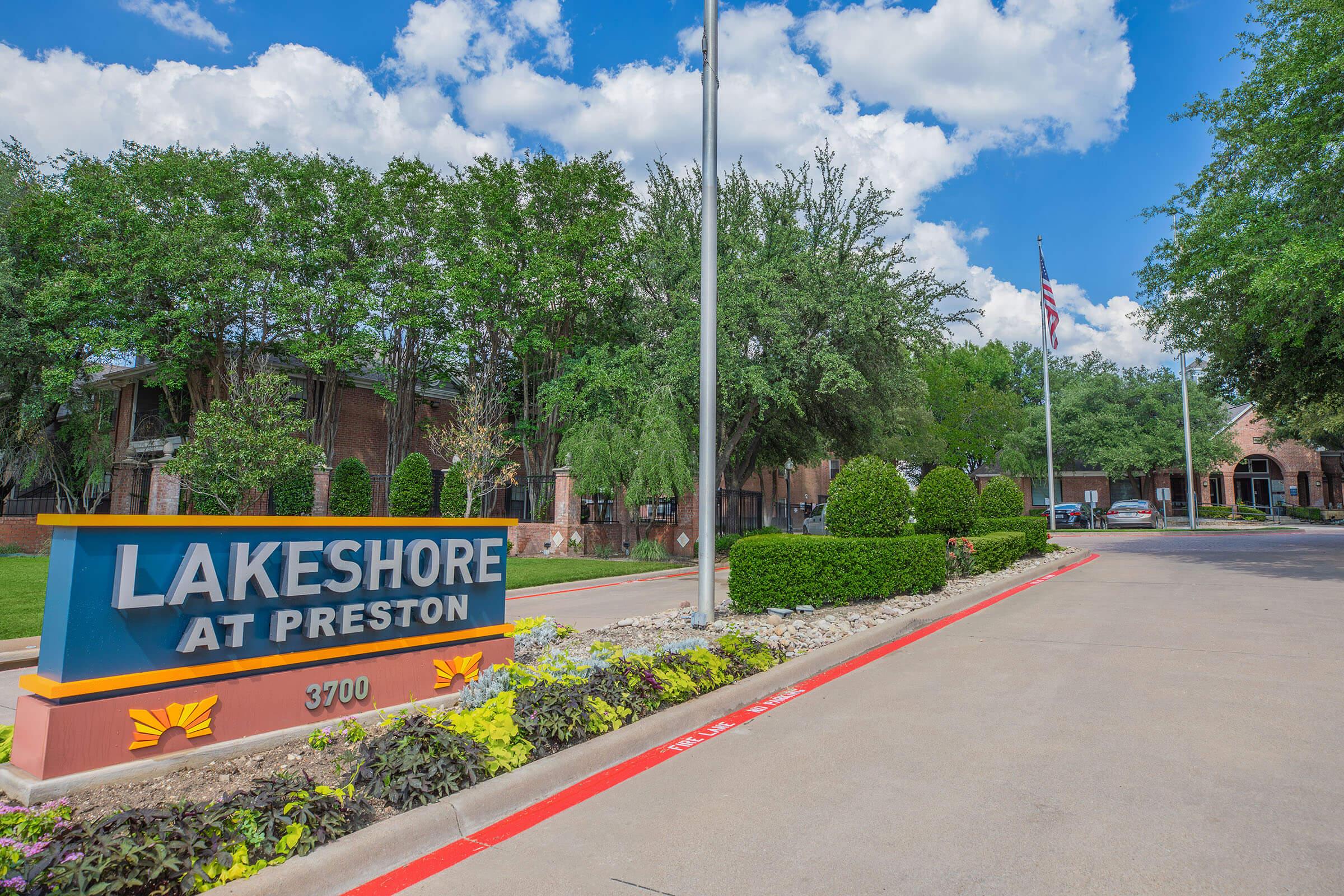 Sign for Lakeshore at Preston, featuring the property name prominently displayed. Surrounding landscape includes neatly trimmed bushes and trees under a clear blue sky with clouds. The entrance road is lined with decorative plants and leads to the residential buildings in the background. An American flag is visible nearby.