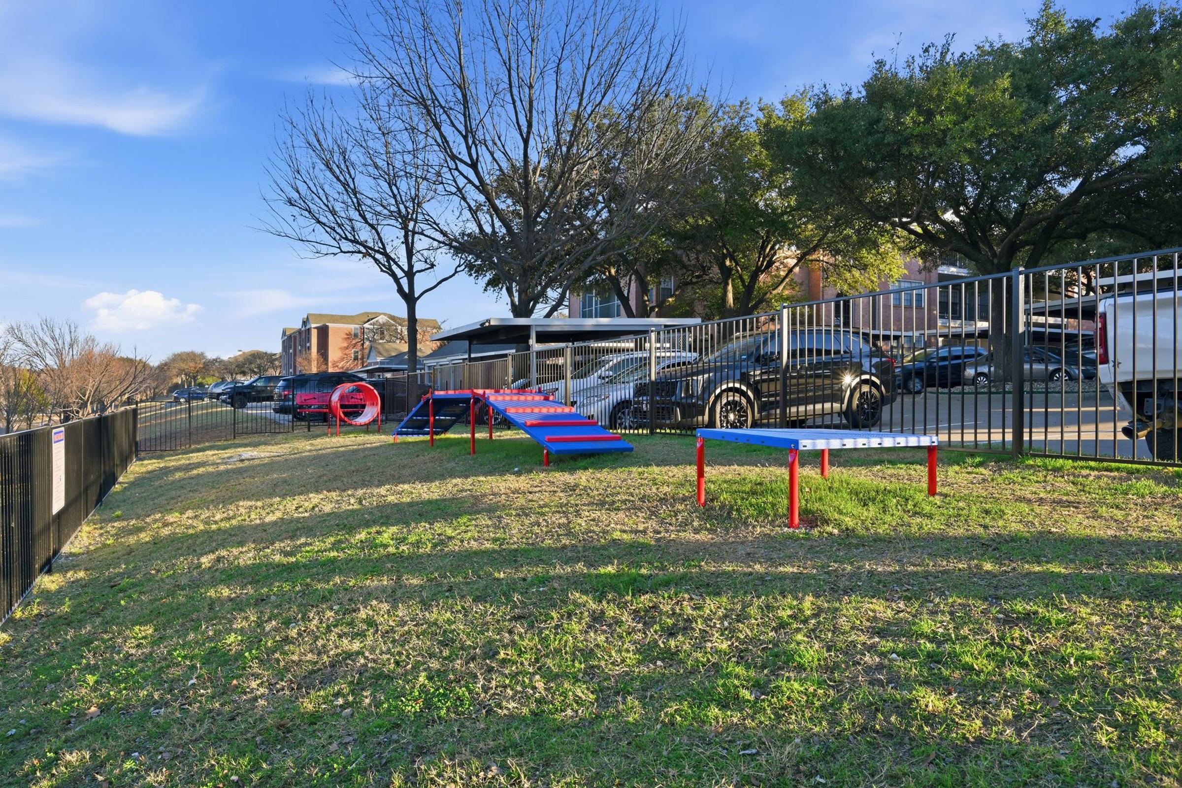 A grassy area with colorful play equipment, including a set of ramps and a circular tunnel, surrounded by a black fence. Nearby vehicles are parked along the road, with trees in the background and a blue sky above.
