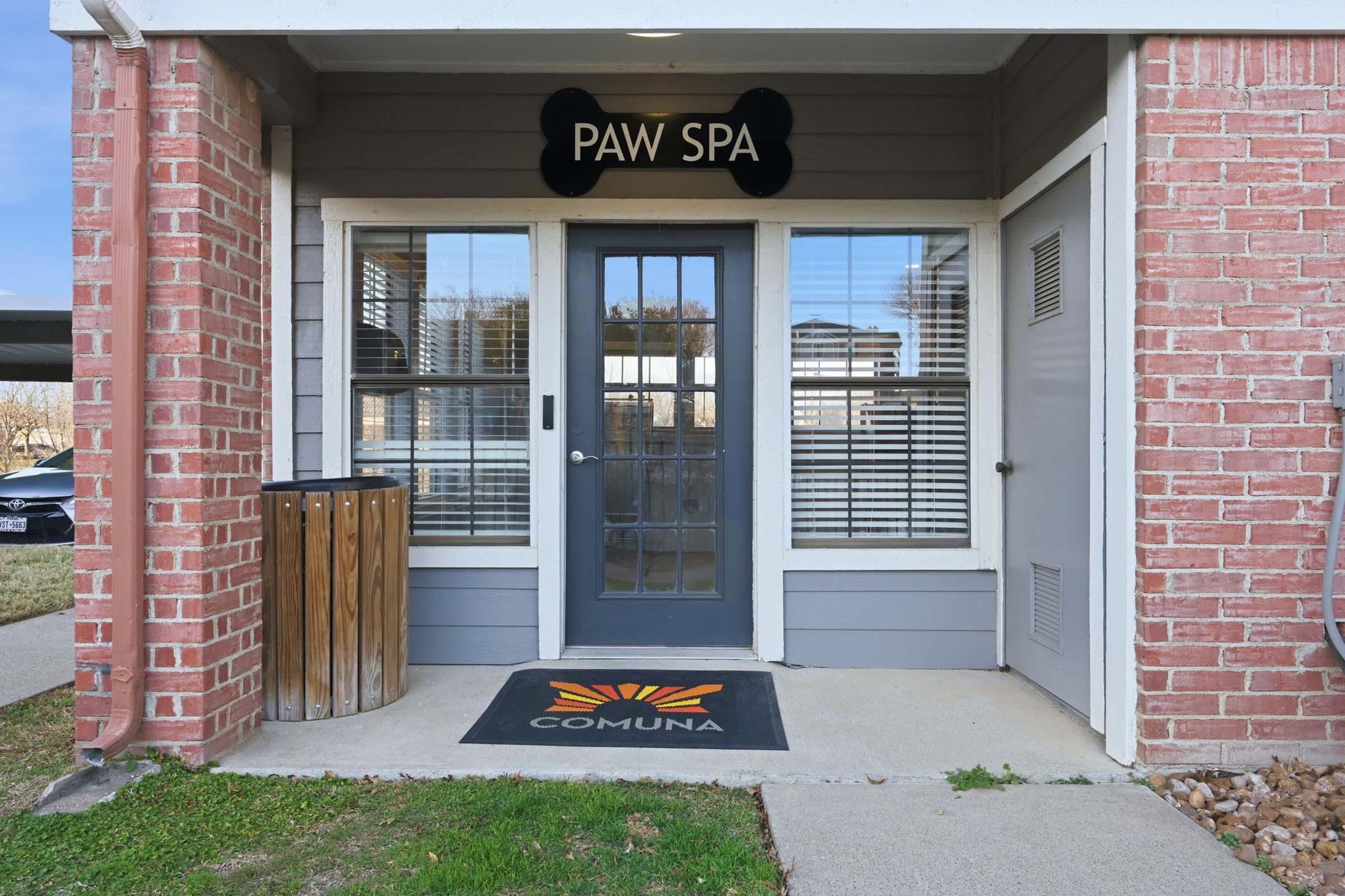 Entrance of a pet grooming facility named "Paw Spa." The building features a welcoming door with glass panels, a decorative sign above, and a "COMUNA" doormat. To the side, there is a trash bin, and the area is surrounded by grass and concrete pathways.