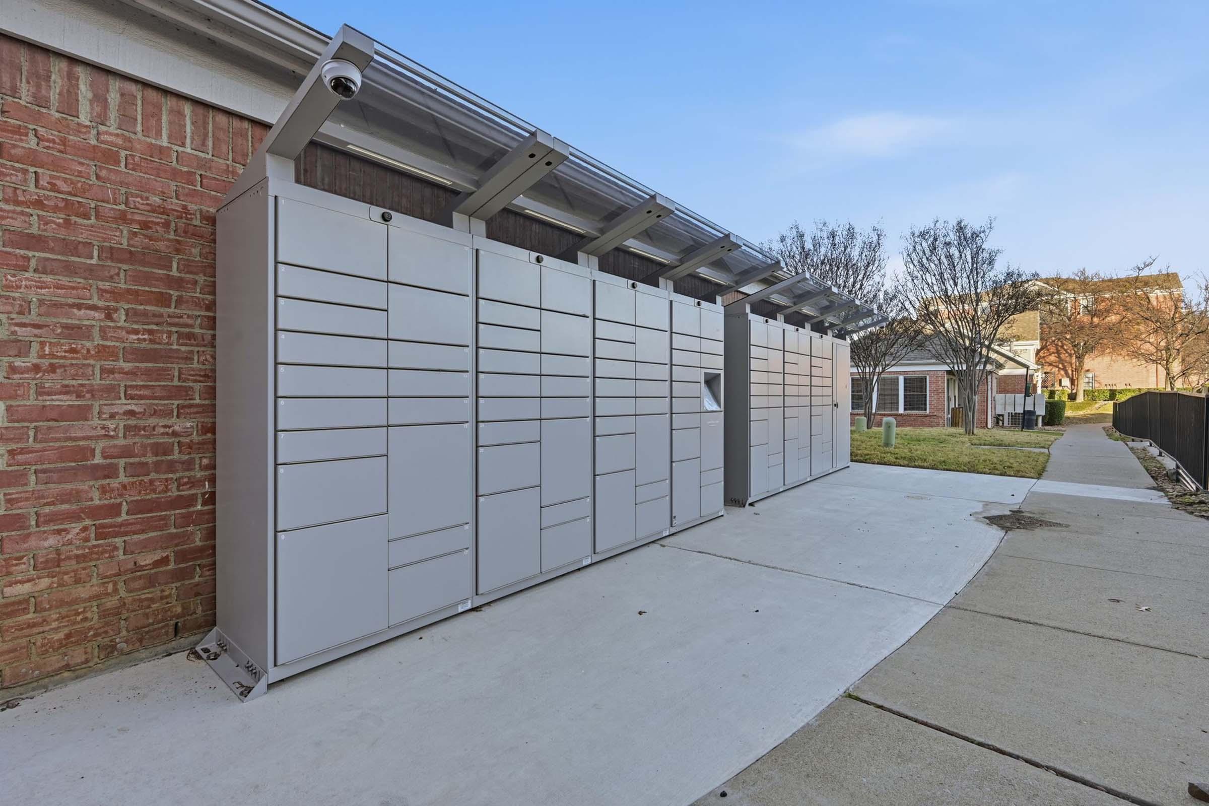 A row of metallic package lockers arranged against a brick wall. The lockers are neatly aligned and feature various compartments for secure mail delivery. The surrounding area includes a paved walkway and grass, with trees in the background under a clear blue sky.