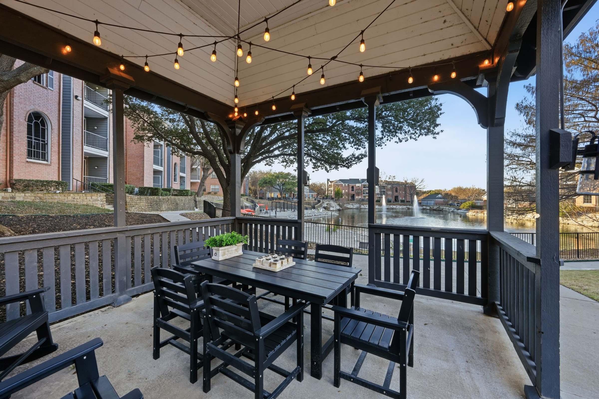 A cozy outdoor gazebo featuring a wooden dining table and chairs, illuminated by string lights. In the background, a serene view of a lake and residential buildings. The area is surrounded by trees, creating a peaceful atmosphere for relaxation and gatherings.