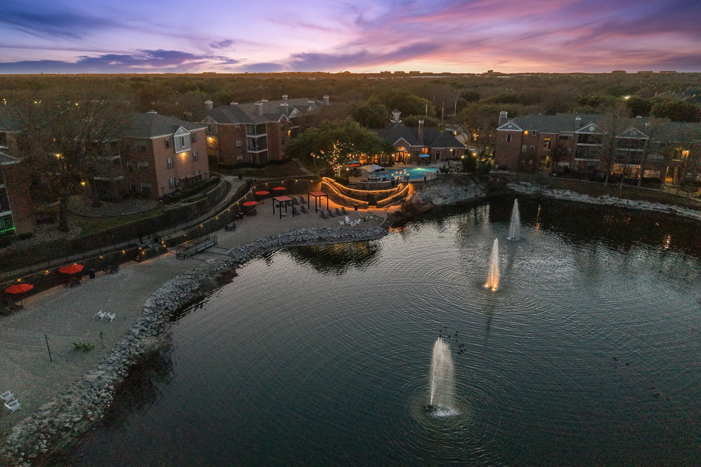 A serene waterfront view at dusk featuring a peaceful lake surrounded by residential buildings. Illuminated fountains spout water into the lake, and outdoor seating areas with umbrellas are visible along the shore. The sky transitions from sunset to twilight, adding vibrant colors to the scene.