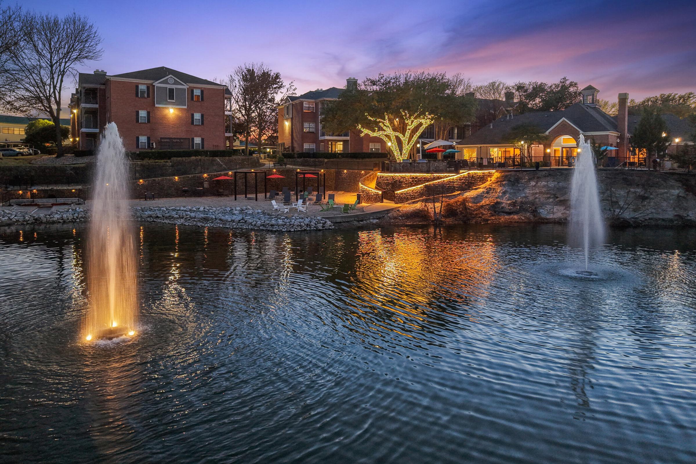 A tranquil lake scene at dusk, featuring two illuminated fountains spraying water. Surrounding the lake are buildings with warm lighting, trees, and seating areas. The sky transitions from blue to purple, reflecting in the water, creating a serene atmosphere.