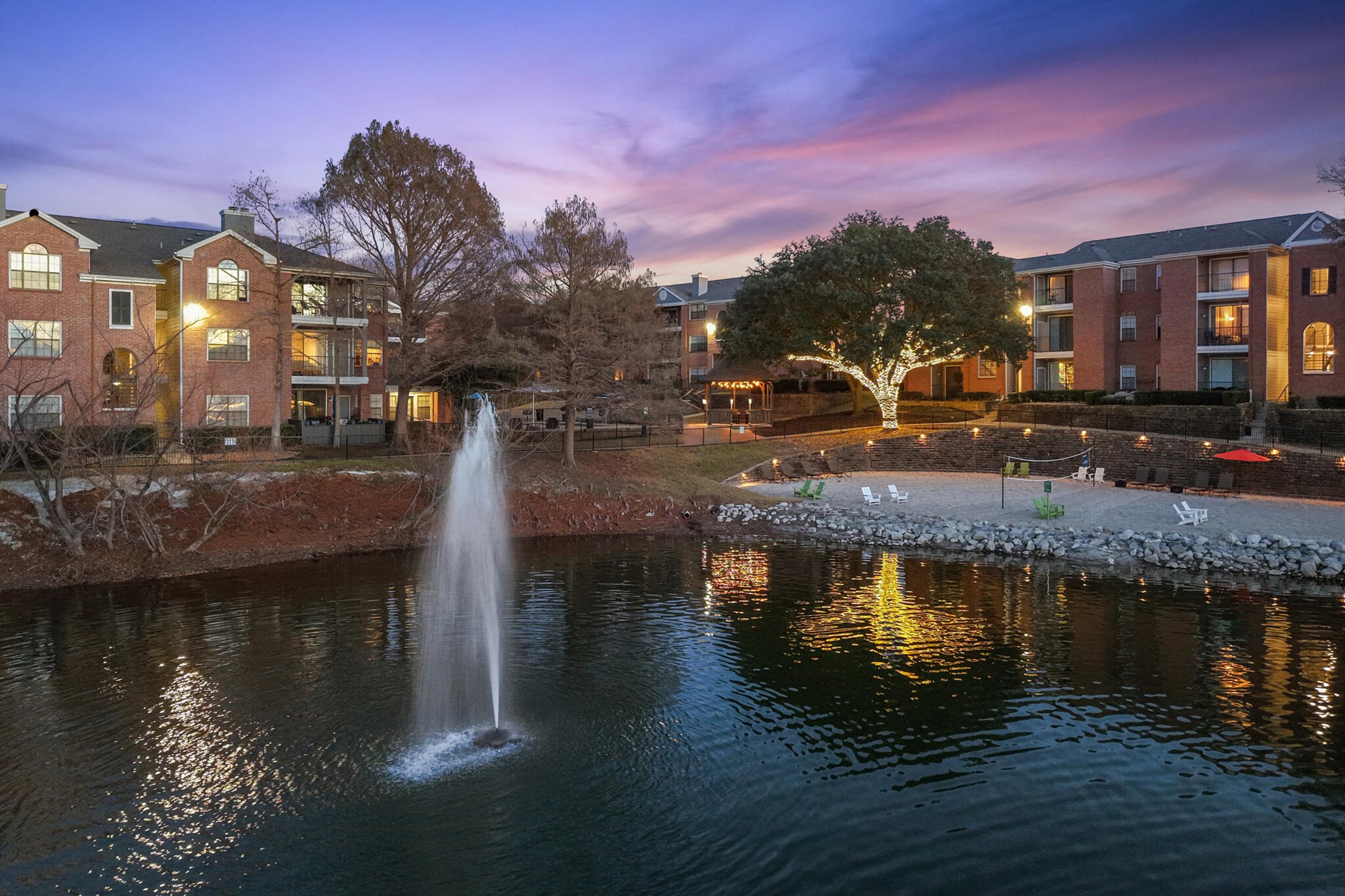 A serene view of a small lake featuring a fountain, surrounded by residential buildings. The sky glows with pastel colors during sunset, reflecting on the water's surface. Trees and chairs are visible along the shore, creating a peaceful atmosphere for relaxation and enjoyment.