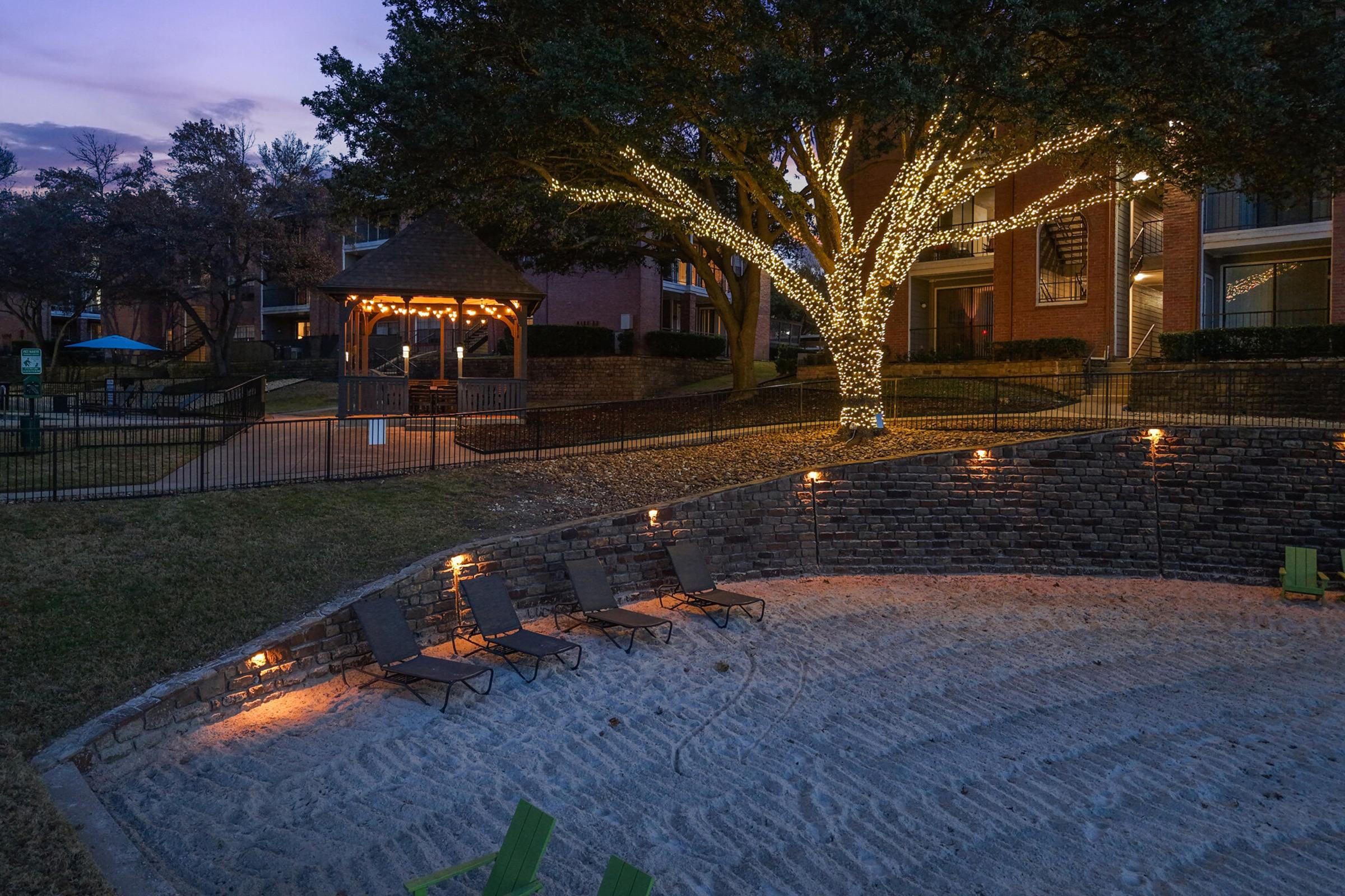 A serene outdoor area with a sand pit, three lounge chairs, and a gazebo adorned with lights. A large, lit tree stands nearby, surrounded by grass and brick walls. The evening sky hints at twilight, enhancing the calm ambiance of the space.