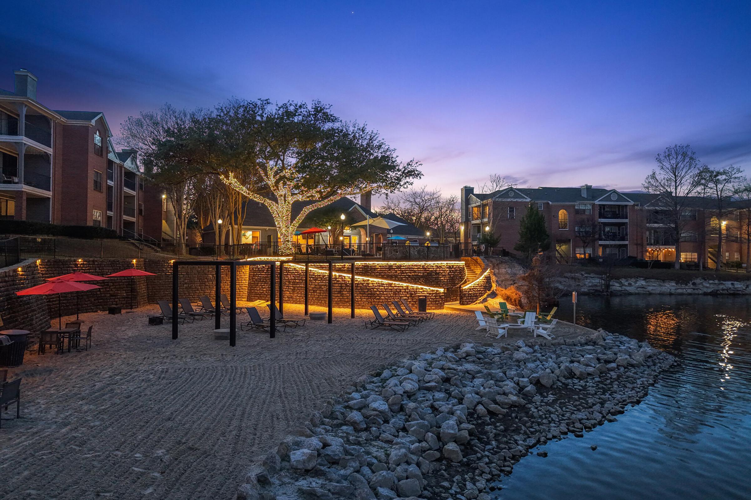 A tranquil lakeside scene at dusk featuring a sandy beach area with lounge chairs and red umbrellas. The shoreline is lined with rocks, and there are illuminated trees nearby. In the background, elegant apartment buildings are visible, reflecting the soft colors of the evening sky.