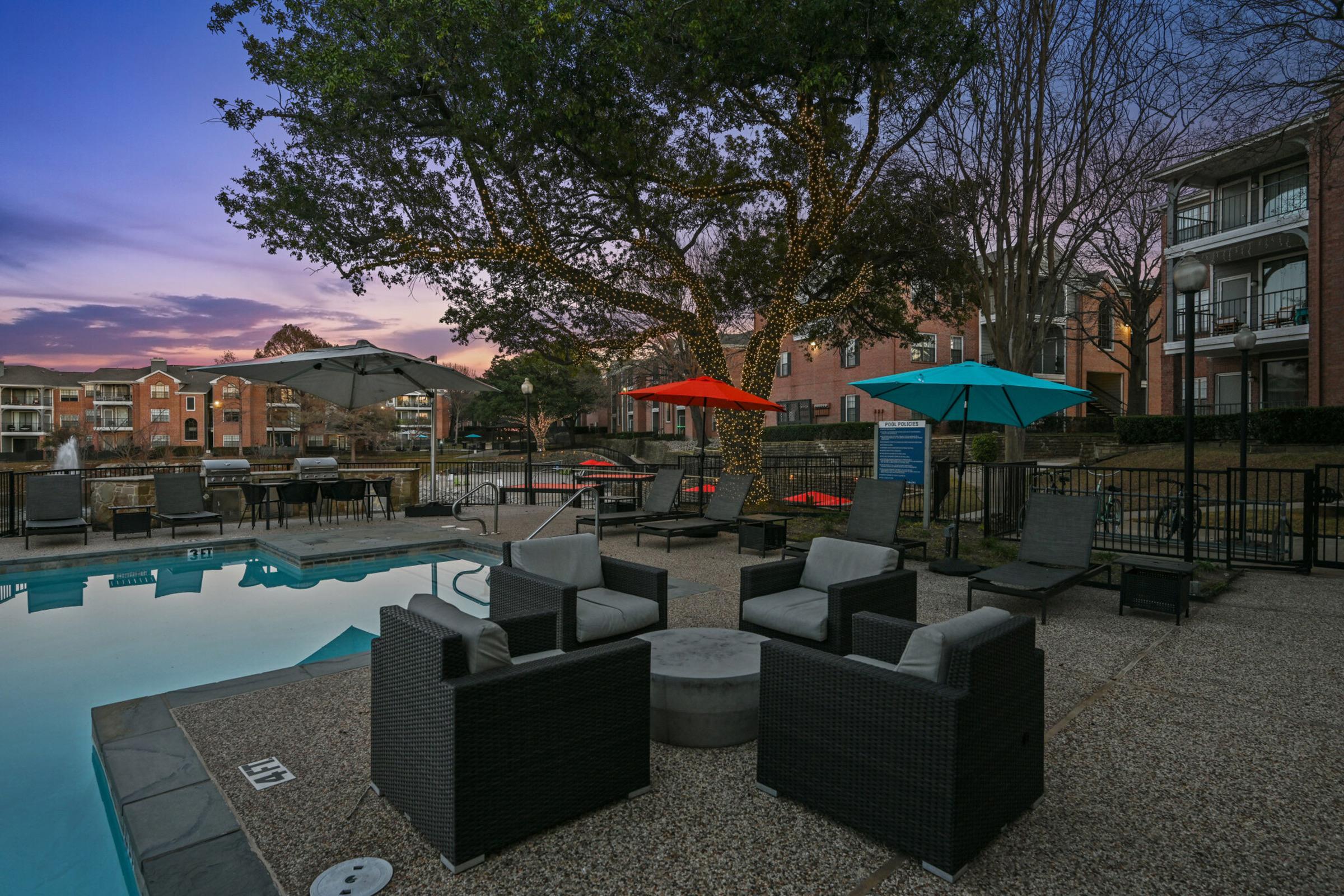 Outdoor pool area at dusk, featuring modern seating arrangements around a cozy fire pit. Colorful umbrellas provide shade, while the surrounding apartment buildings are softly lit. Trees in the background add to the serene atmosphere, enhancing the inviting ambiance of the space.