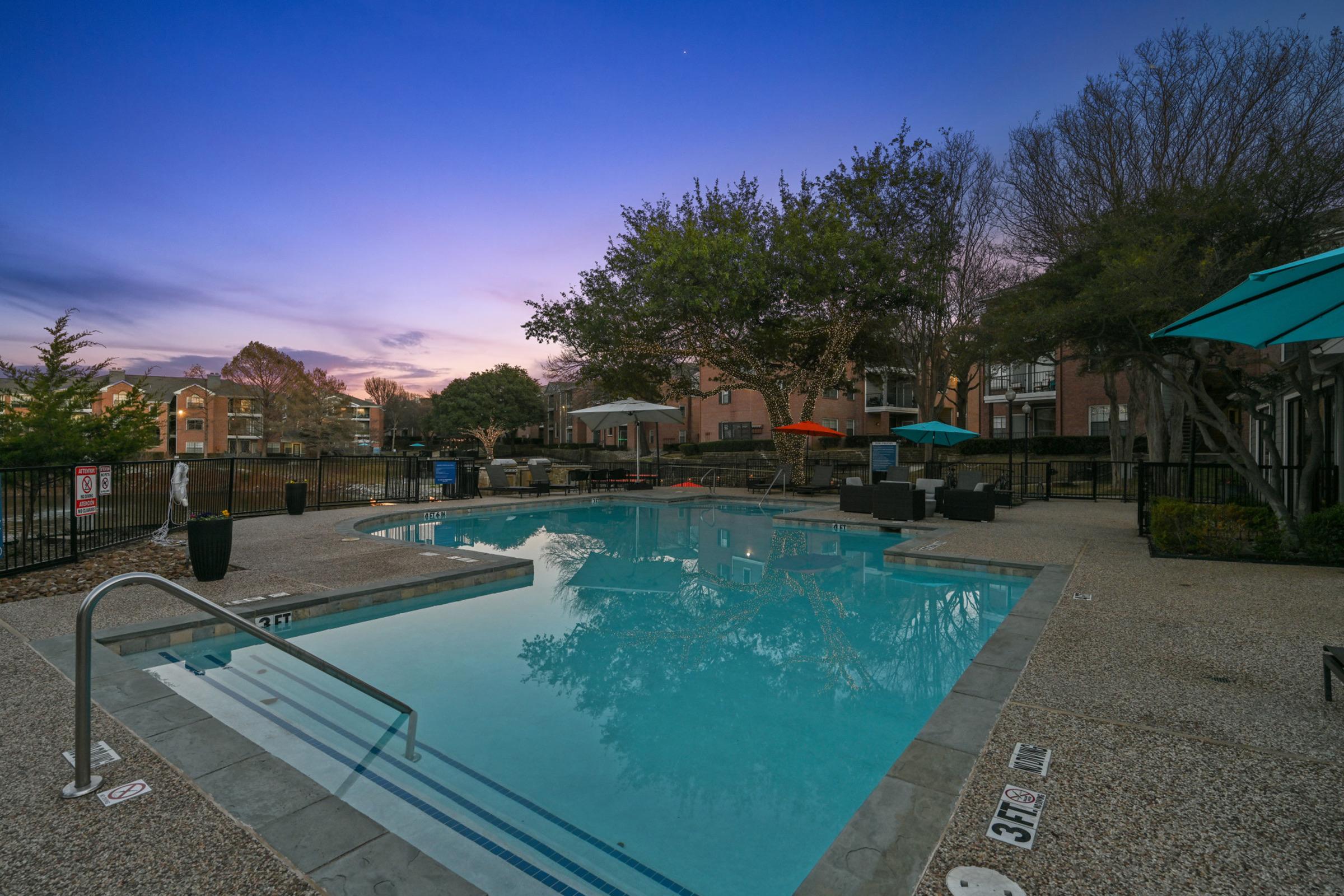 A tranquil pool area at dusk, featuring a clear water pool with steps leading in, surrounded by lounge chairs and colorful umbrellas. Lush trees and modern buildings are visible in the background, creating a peaceful ambiance.