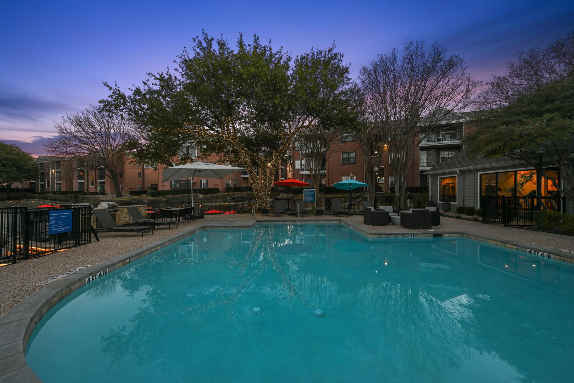 A tranquil evening scene of a pool area surrounded by trees and residential buildings. The water reflects the sky at dusk, with several lounge chairs and umbrellas in bright colors. Soft lighting creates a cozy atmosphere, inviting relaxation and leisure in the outdoor space.