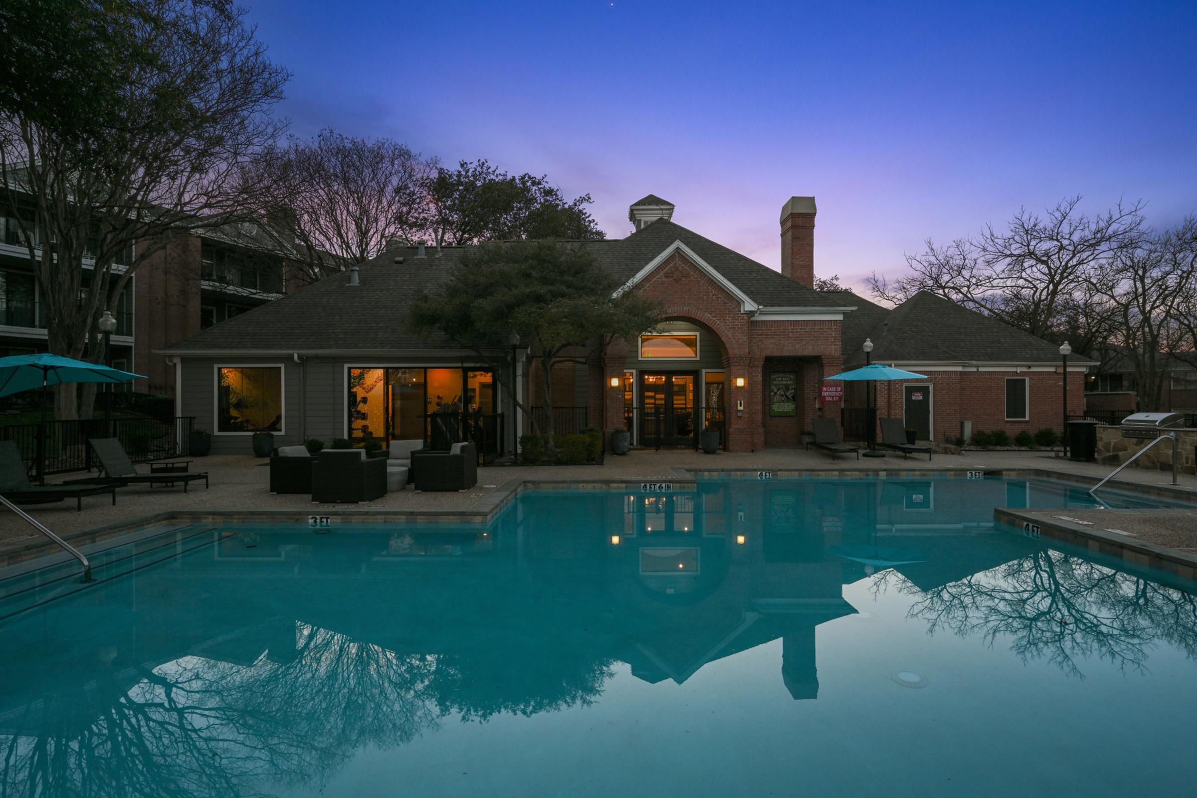 A serene evening view of a pool area with lounge chairs and umbrellas. The reflection of a brick building and trees is visible in the water, under a twilight sky. Warm lights glow from the building's windows, creating a welcoming atmosphere.