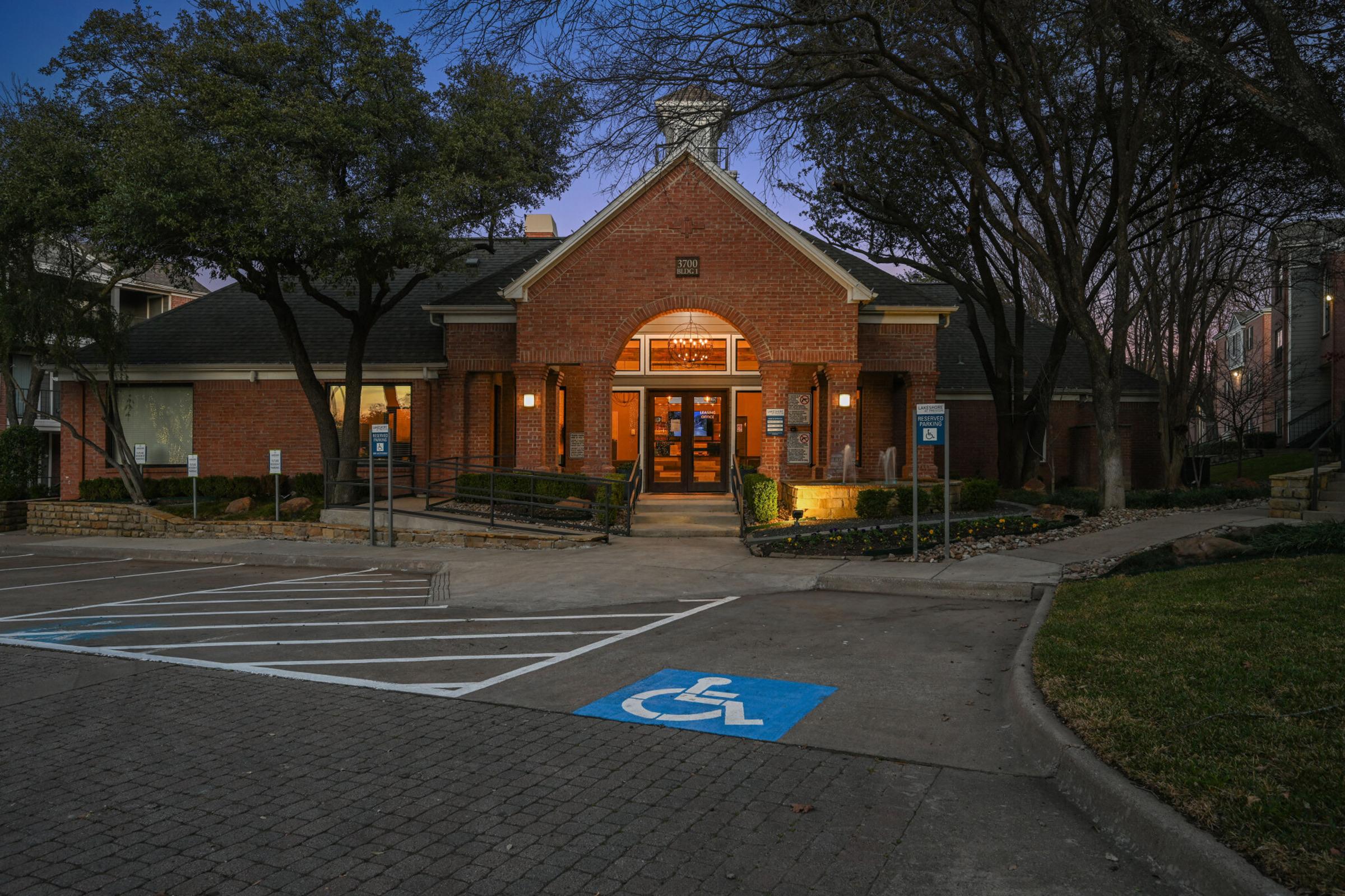 A brick building with a peaked roof and large entrance illuminated by warm light, surrounded by trees and landscaping. A handicap parking space is marked in blue near the entrance, with a path leading up to the door. The scene is set during twilight with a soft glow in the sky.