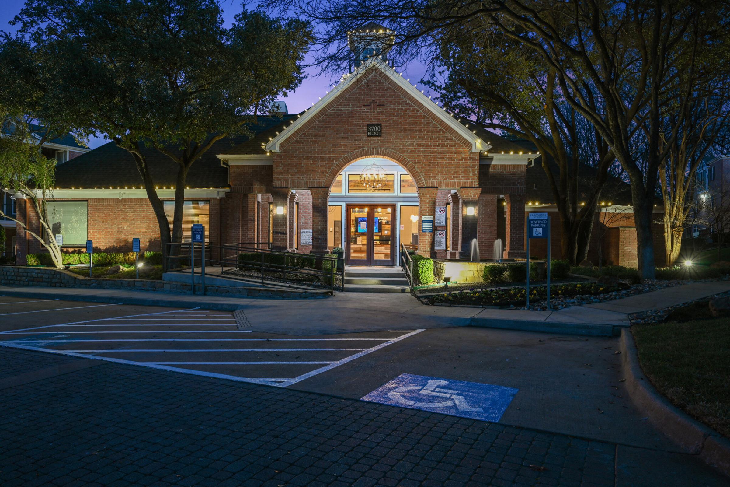 A brick building with a peaked roof, lit warmly from within, surrounded by trees. The entrance features double doors and a welcoming sign. A blue wheelchair-accessible parking spot is visible in the foreground, alongside marked parking spaces. The scene is set during twilight, with soft lighting accentuating the architecture.