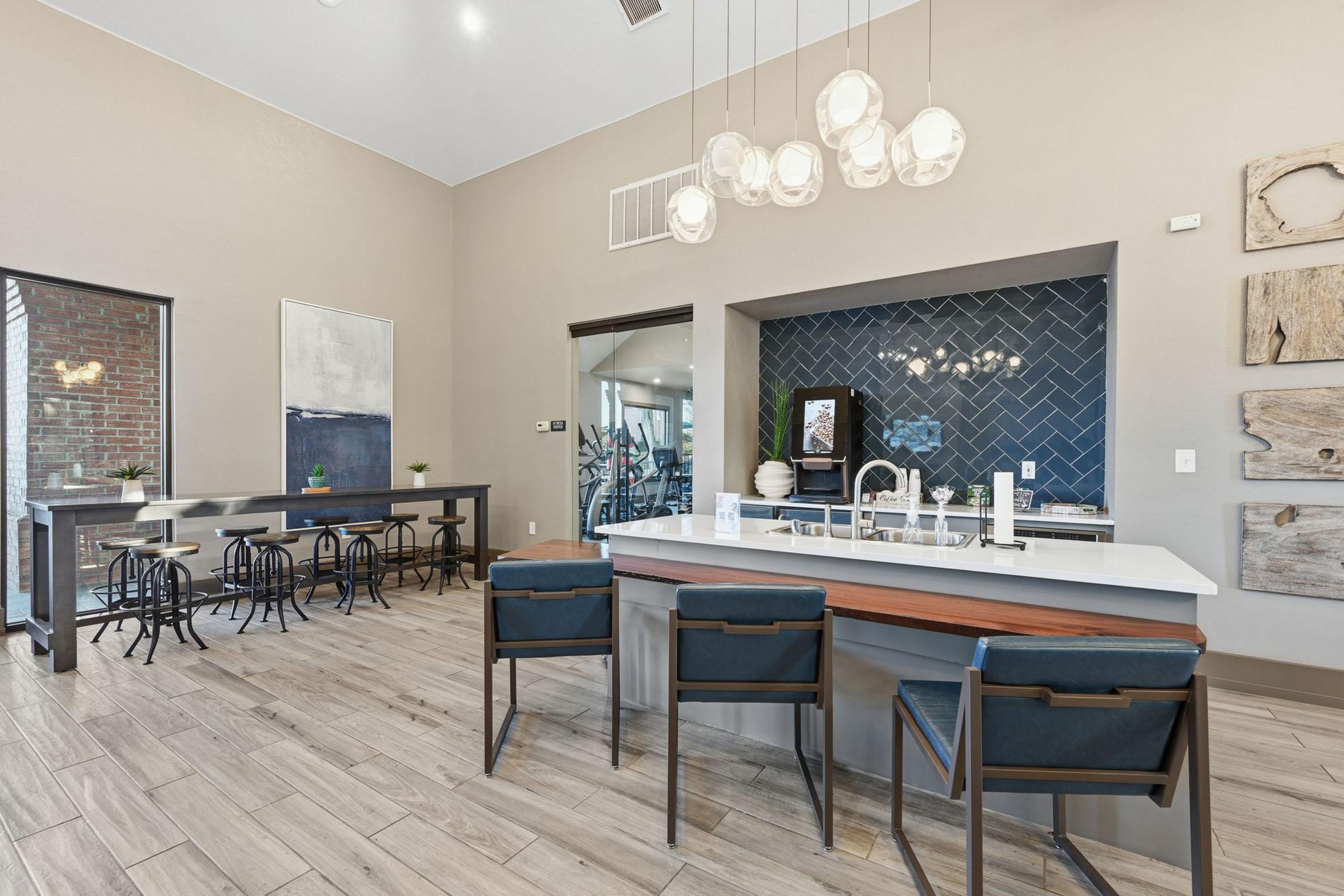 Interior of a modern lounge area featuring a sleek kitchen island with stools, stylish light fixtures, and a textured backsplash. There are small tables and chairs arranged on one side, with large windows providing natural light. The decor includes artwork and wooden accents for a contemporary ambiance.