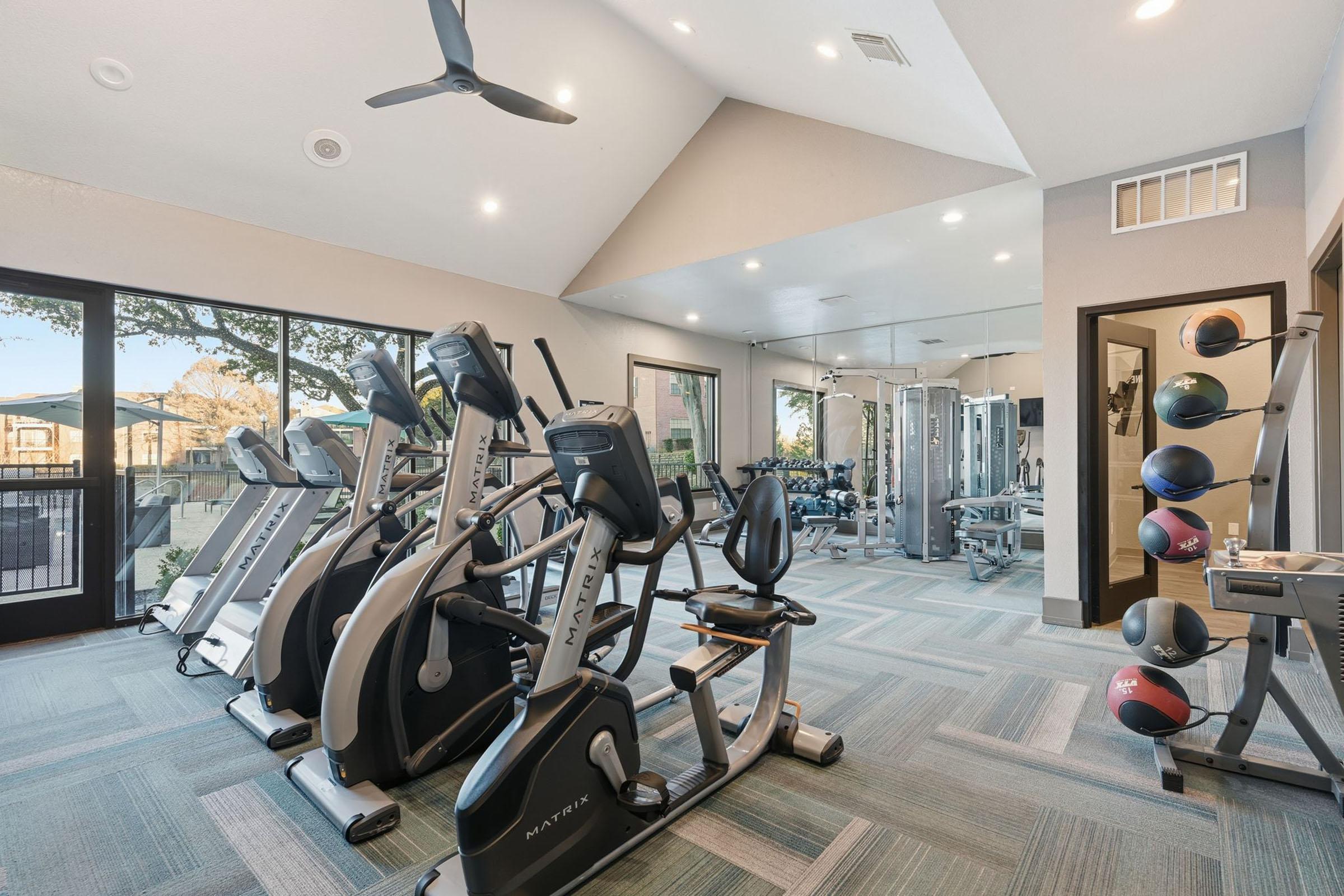 A modern gym interior featuring various exercise machines, including stationary bikes and elliptical machines. The space has large windows allowing natural light, a mirrored wall, and weight training equipment. Stylish flooring and ceiling fans contribute to a comfortable workout environment.