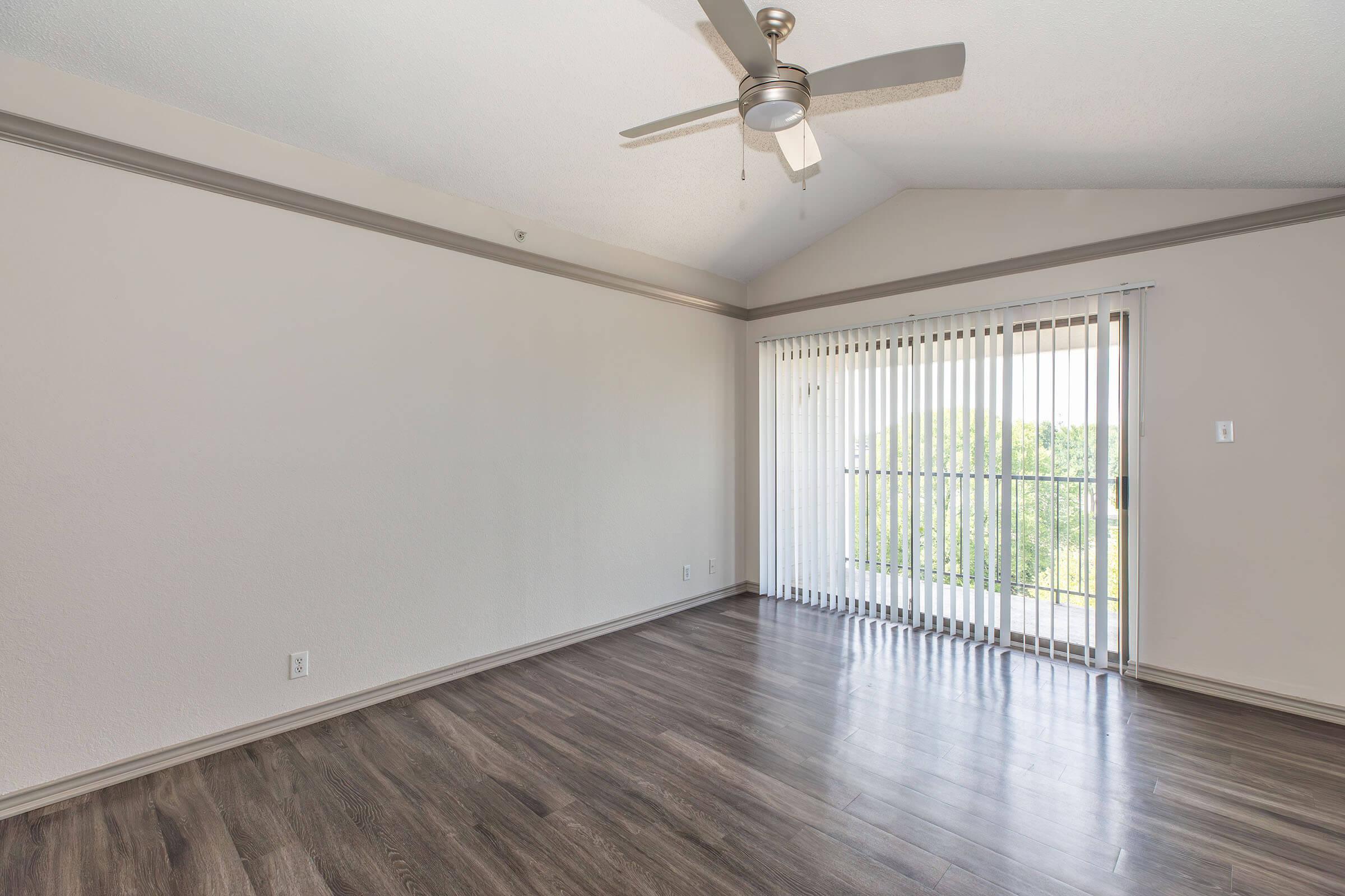 Spacious interior of a room featuring light-colored walls, high vaulted ceiling with a ceiling fan, and wood-like flooring. There are vertical blinds covering a sliding glass door that opens to a balcony overlooking greenery outside. The space is well-lit and uncluttered, providing a fresh and airy atmosphere.