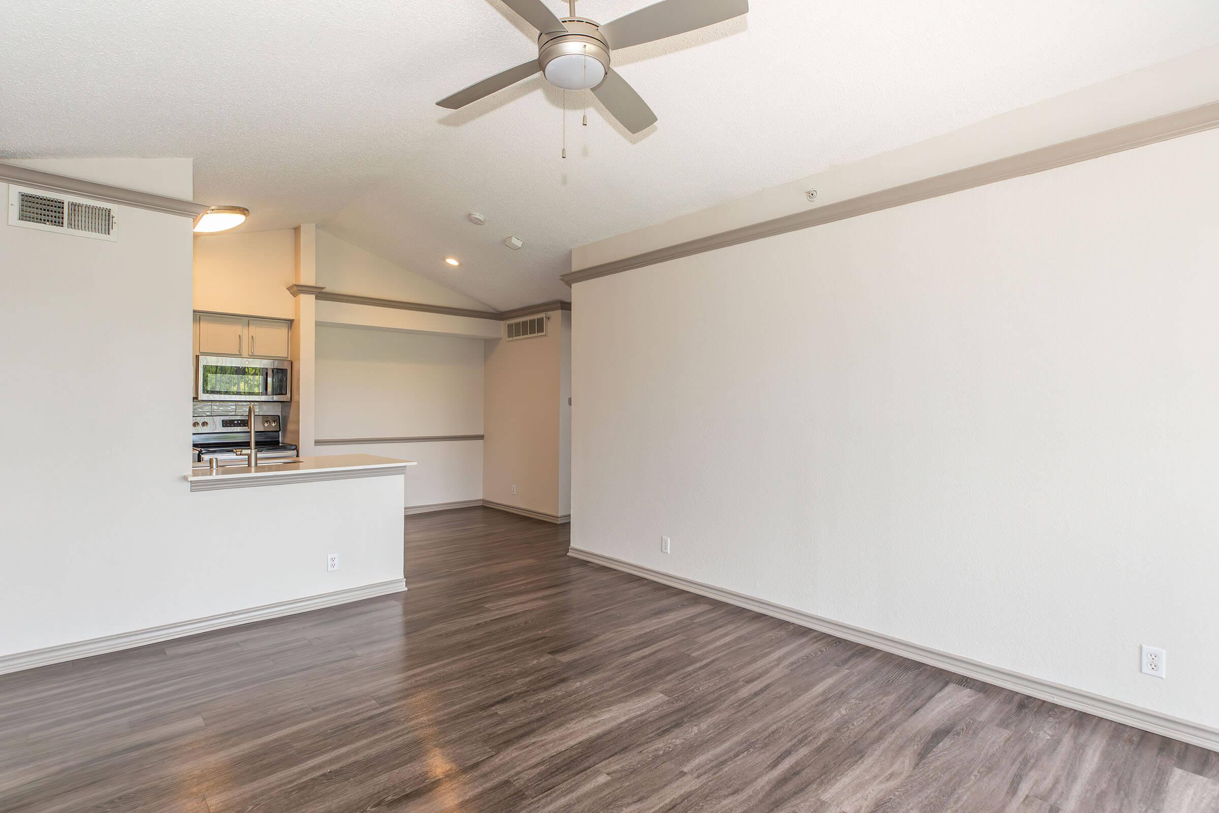 Bright, spacious living area with high ceilings and a ceiling fan. Light-colored walls and wood-like flooring create a modern and airy atmosphere. A small kitchen area is visible in the background, featuring a counter that separates it from the living space. Natural light comes through windows, enhancing the overall openness.