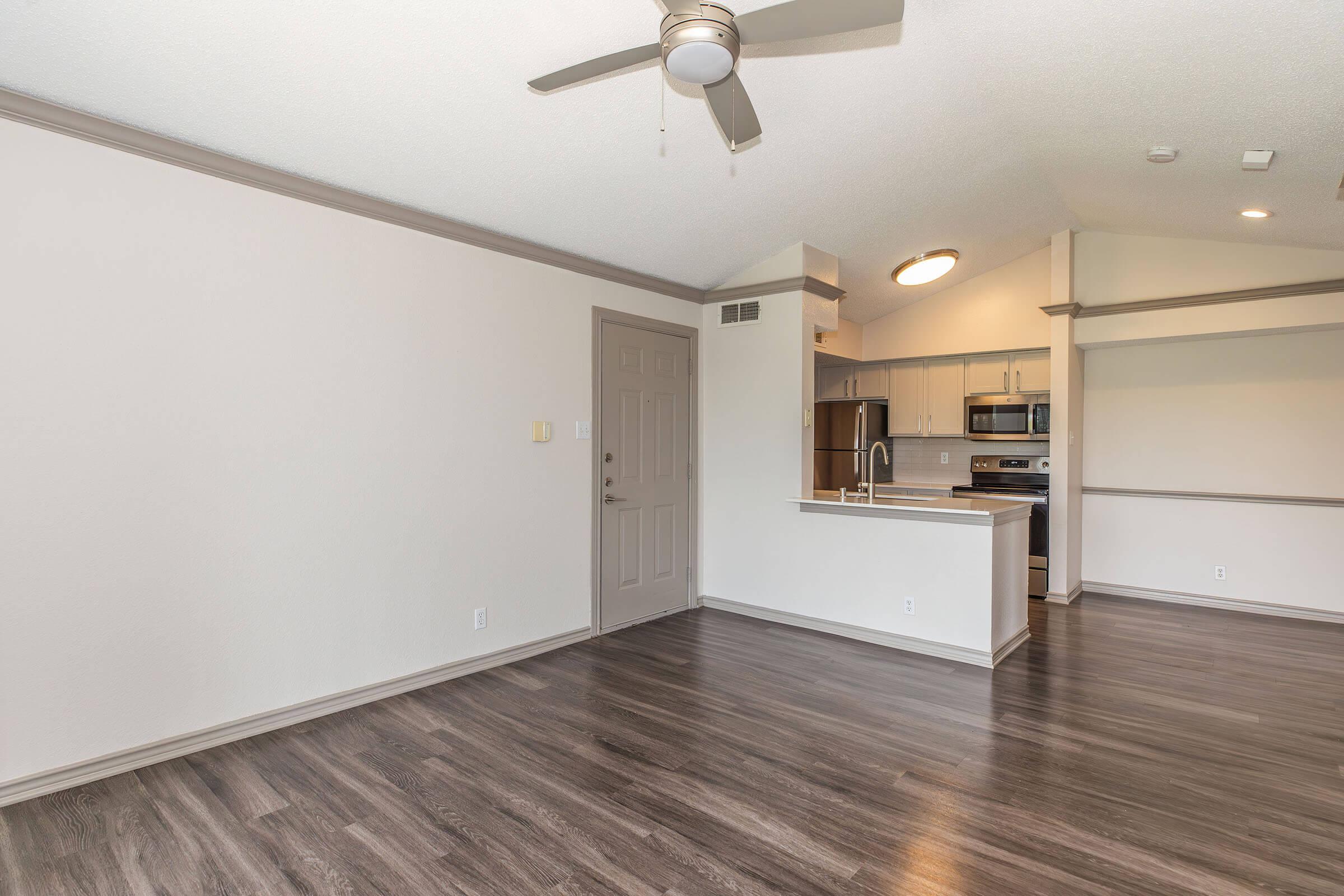 Spacious living area featuring a modern ceiling fan, light-colored walls, and sleek vinyl flooring. An open kitchen with stainless steel appliances is visible in the background, alongside a door leading outside. The design focuses on a clean, airy aesthetic.