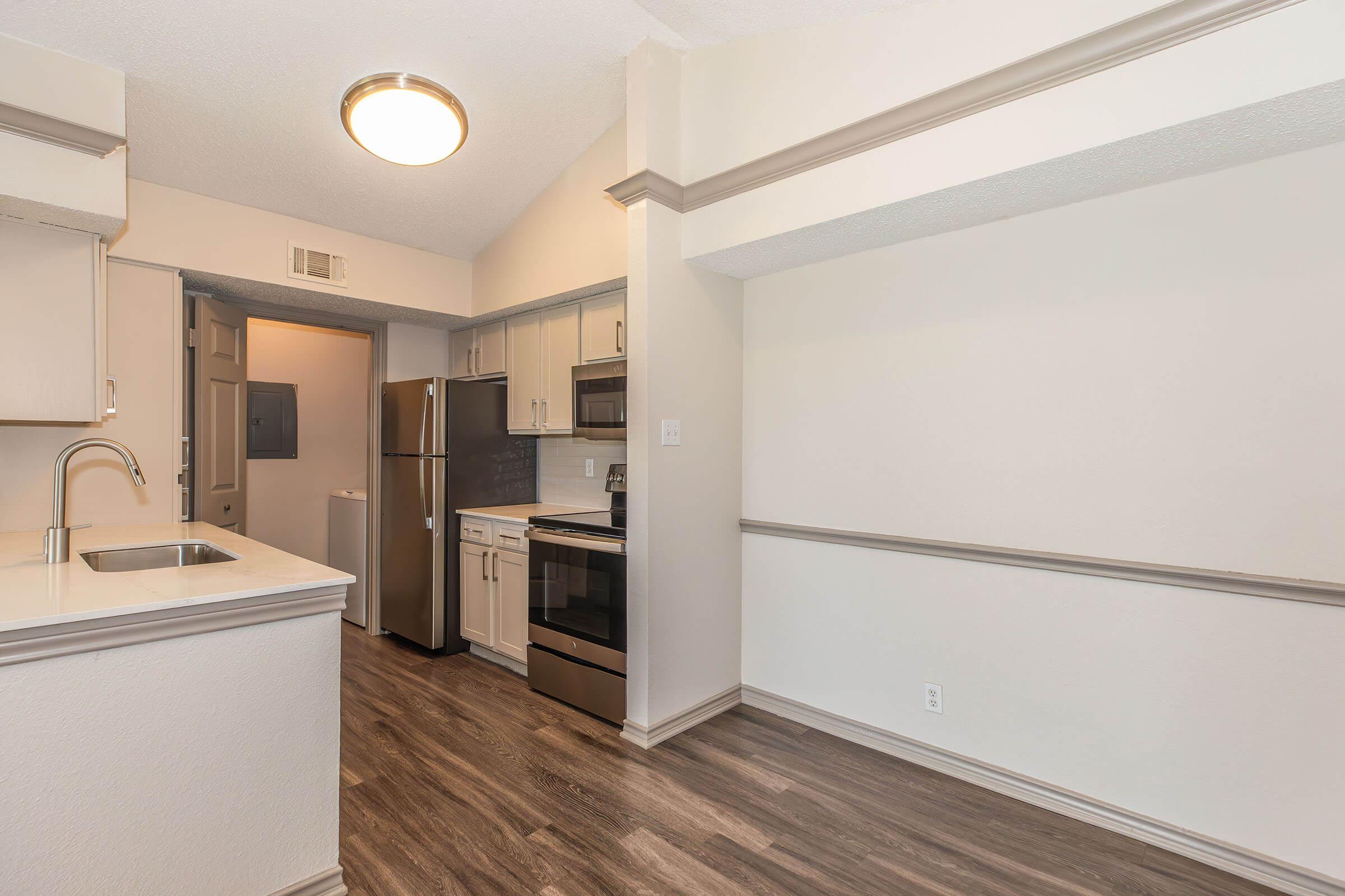 Modern kitchen featuring light-colored cabinets, stainless steel appliances, and a white countertop. The open layout includes a view of a nearby room with a door. The flooring is a warm wood tone, enhancing the spacious feel of the area. Natural light is provided by a ceiling light fixture.