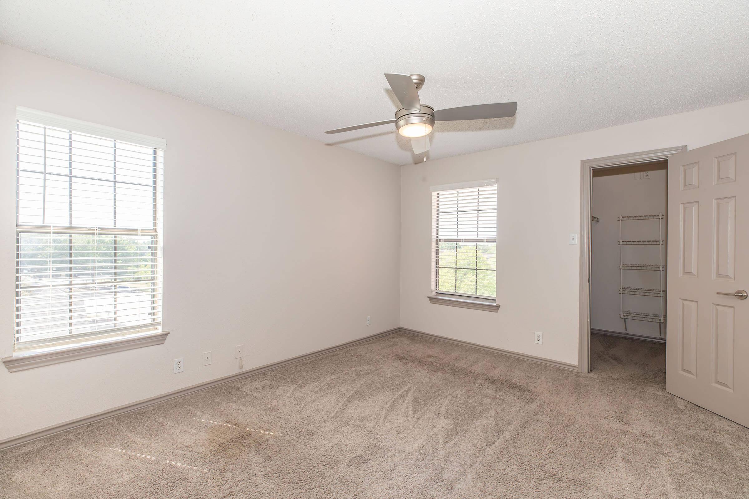 A neutral-toned bedroom featuring carpeted flooring, two windows with blinds allowing natural light, and a ceiling fan. An open closet with shelving is visible to the right, and the walls are painted light colors, creating a spacious and airy feel.