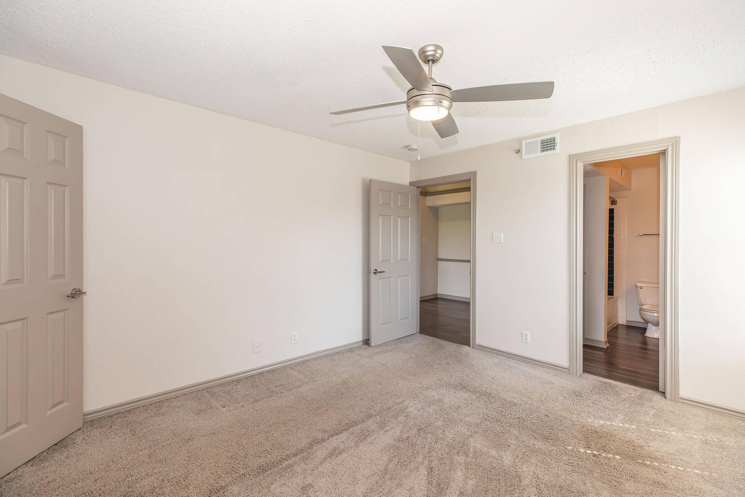 A spacious, neutrally painted bedroom featuring a ceiling fan, plush carpet, and light-colored walls. The room includes two doorways, one leading to a closet and another to an adjacent bathroom. Natural light fills the space, creating a bright and inviting atmosphere.