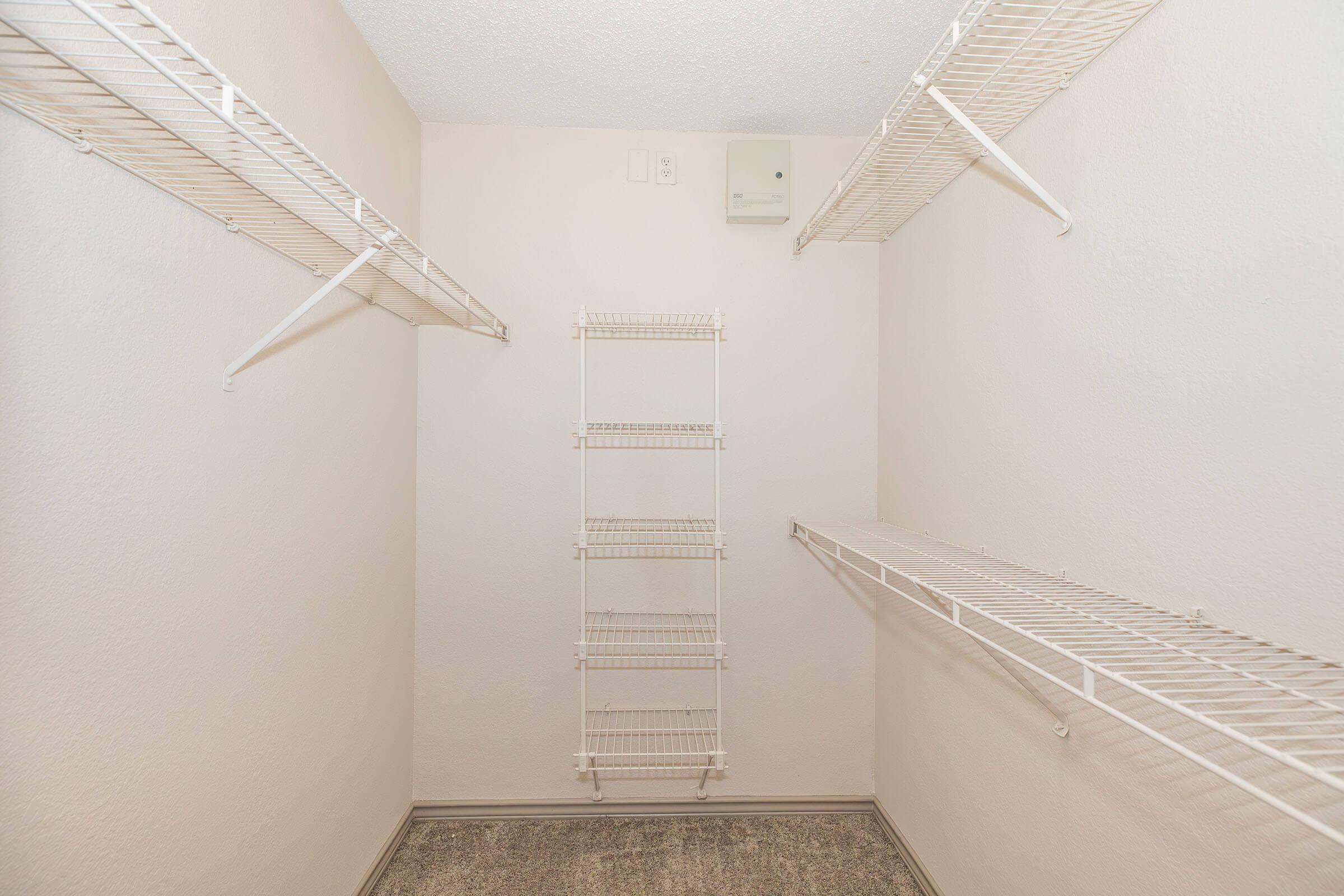 A spacious, empty closet featuring white wire shelving on the walls and a central freestanding rack. The walls are painted a light color, and the floor has a neutral carpet. There are no items or decorations visible in the closet.