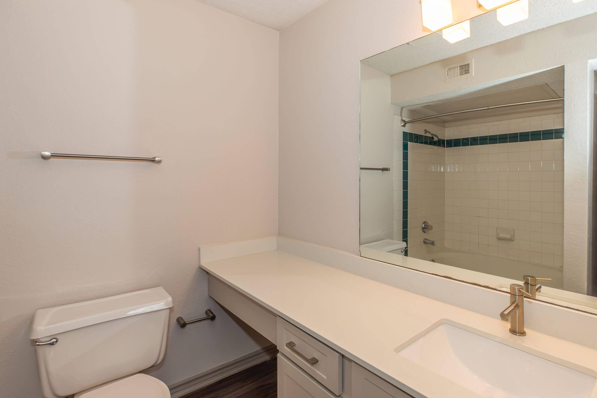 A clean, modern bathroom featuring a white countertop with a sink, a large mirror above, a toilet on the left, and a shower area in the background. The walls are painted light, with a strip of blue tiles lining the shower. Natural light illuminates the space from the ceiling fixtures.
