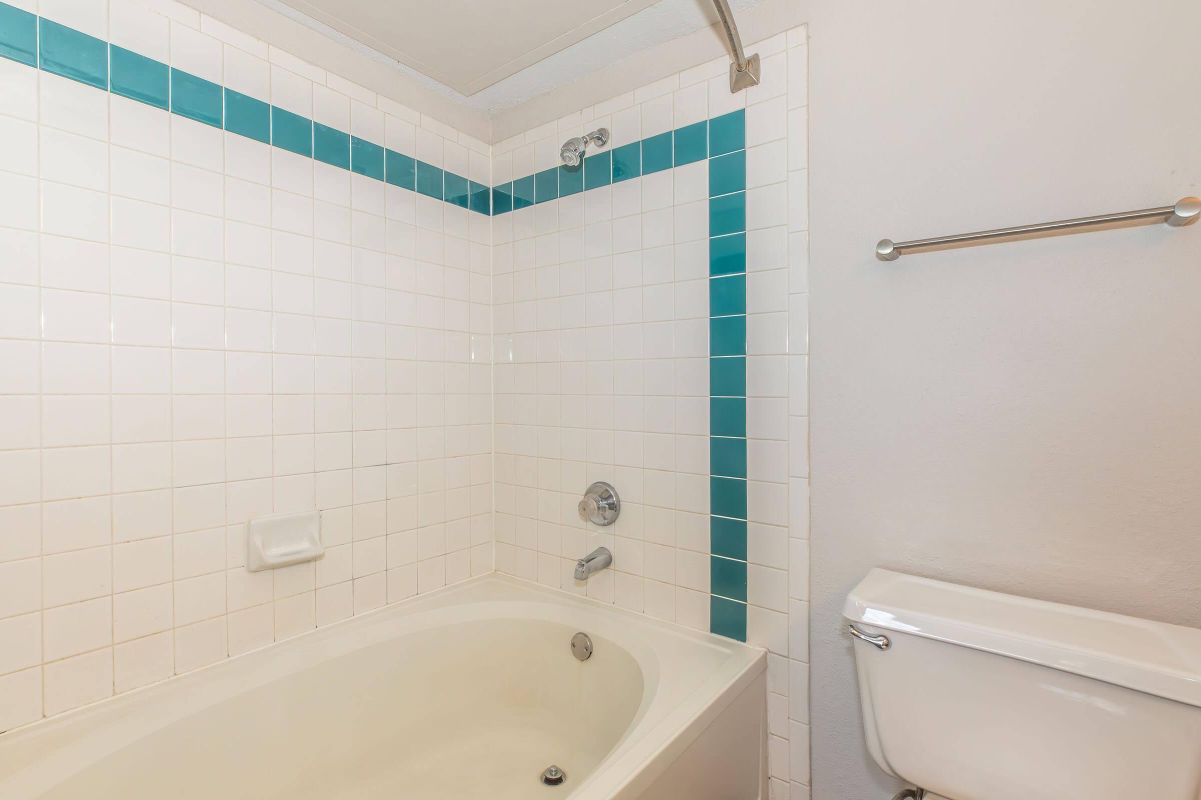 A clean and simple bathroom featuring a white bathtub with a showerhead, white tiled walls with teal accents, and a toilet. The light colors create a bright atmosphere, while the minimalistic design emphasizes functionality.