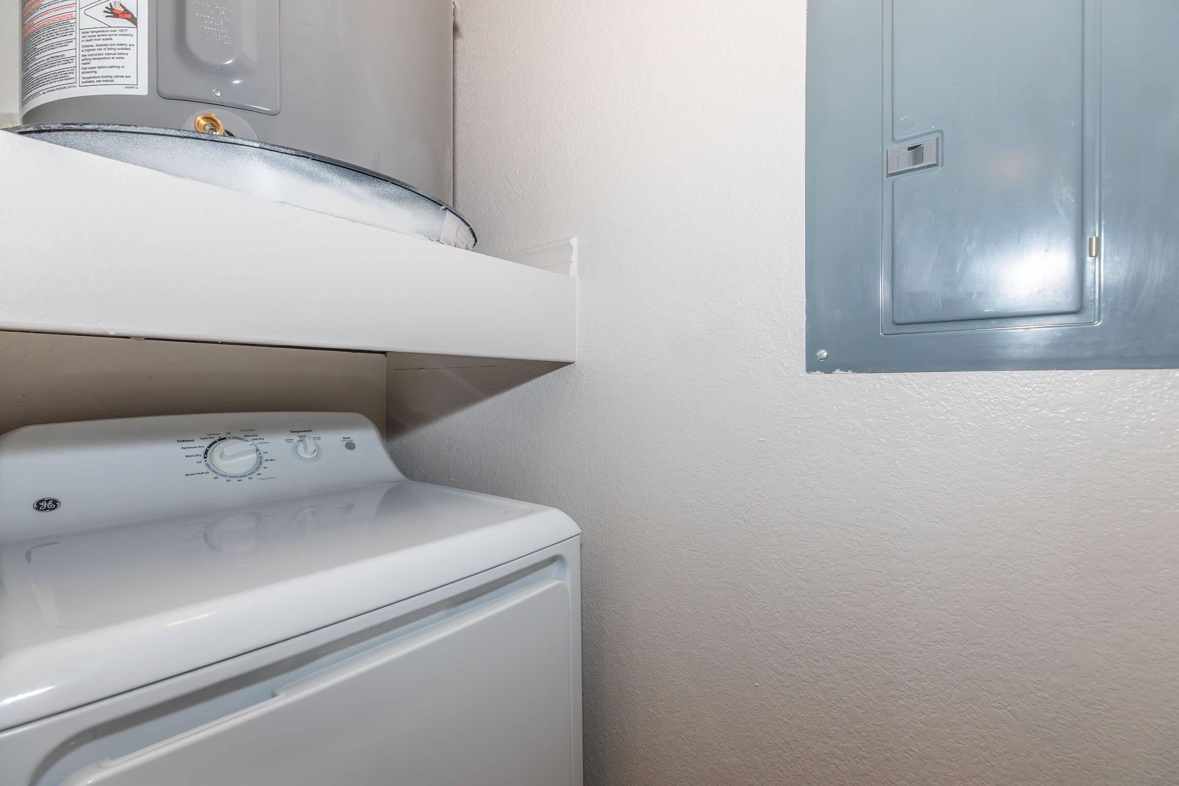 A laundry room featuring a white washer and dryer unit, with a water heater visible above on a shelf. A gray electrical panel is mounted on the wall to the right. The walls are painted a light color, contributing to a clean and functional appearance.
